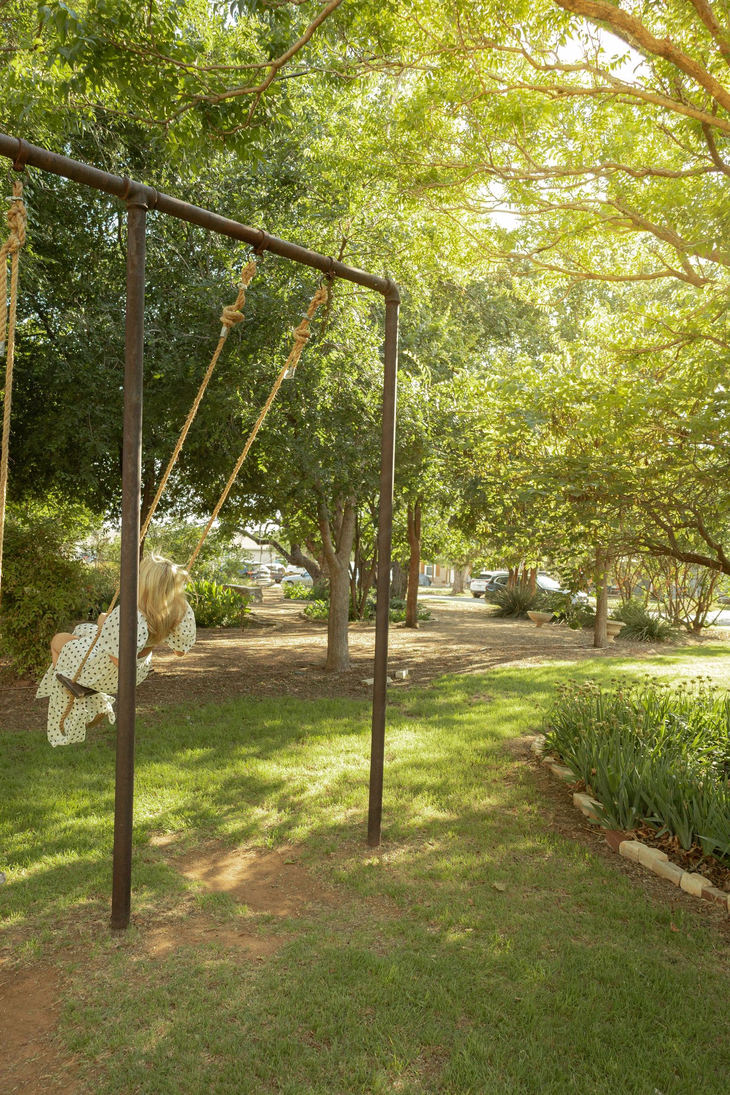 A rustic metal swing set with rope swings in a lush green park surrounded by tall shady trees on a sunny day.