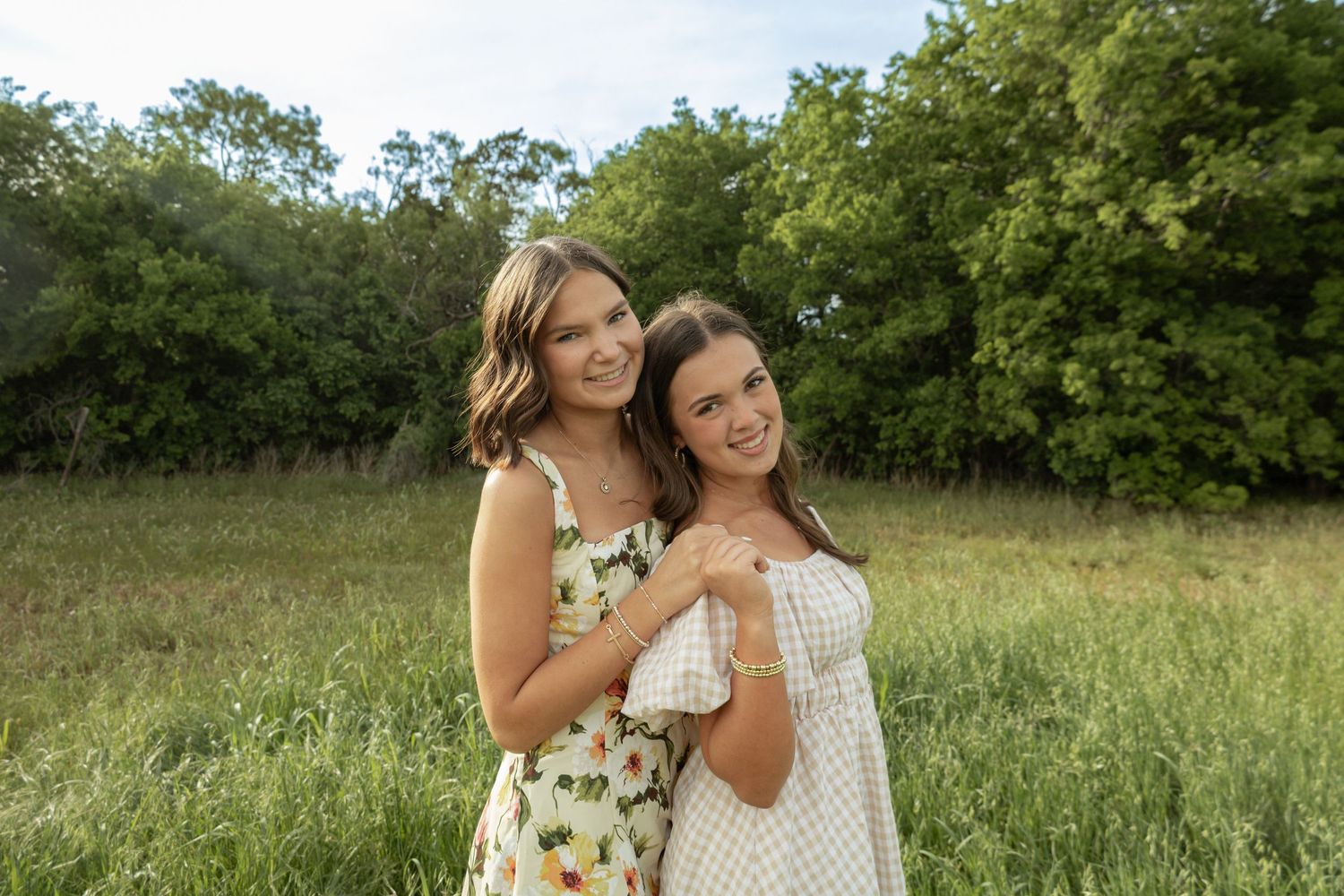 Two young women in floral dresses smiling and embracing in a lush green meadow with trees in the background.