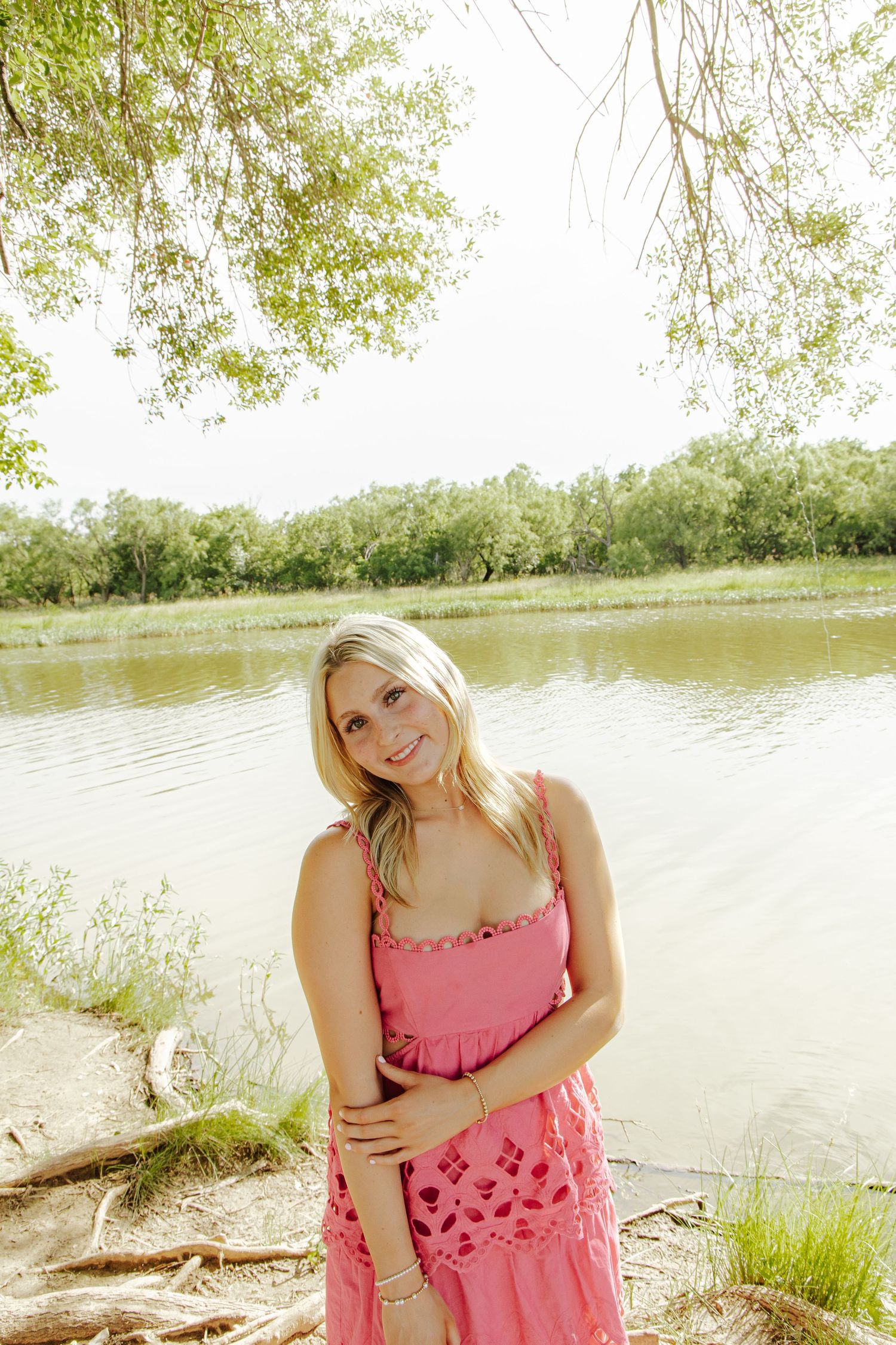 Smiling blonde woman in pink sundress standing by a serene river surrounded by lush green trees.