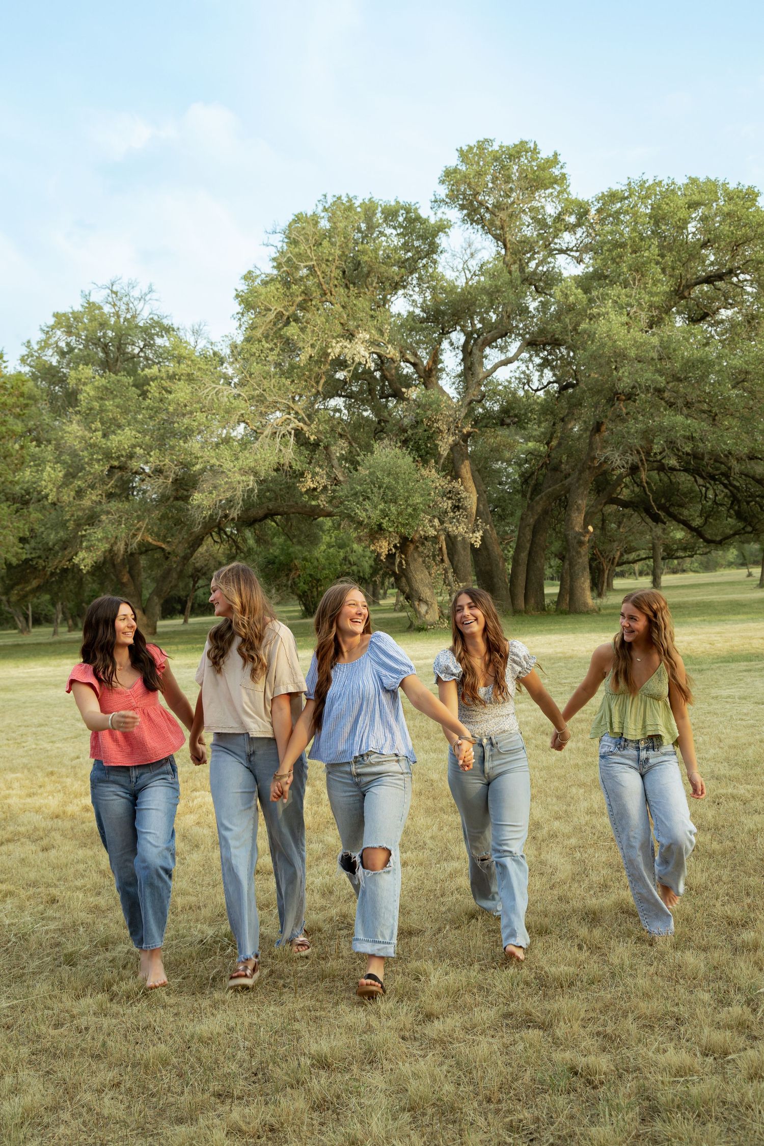 Five young women holding hands walk through a lush green park, wearing casual jeans and colorful tops, smiling joyfully.