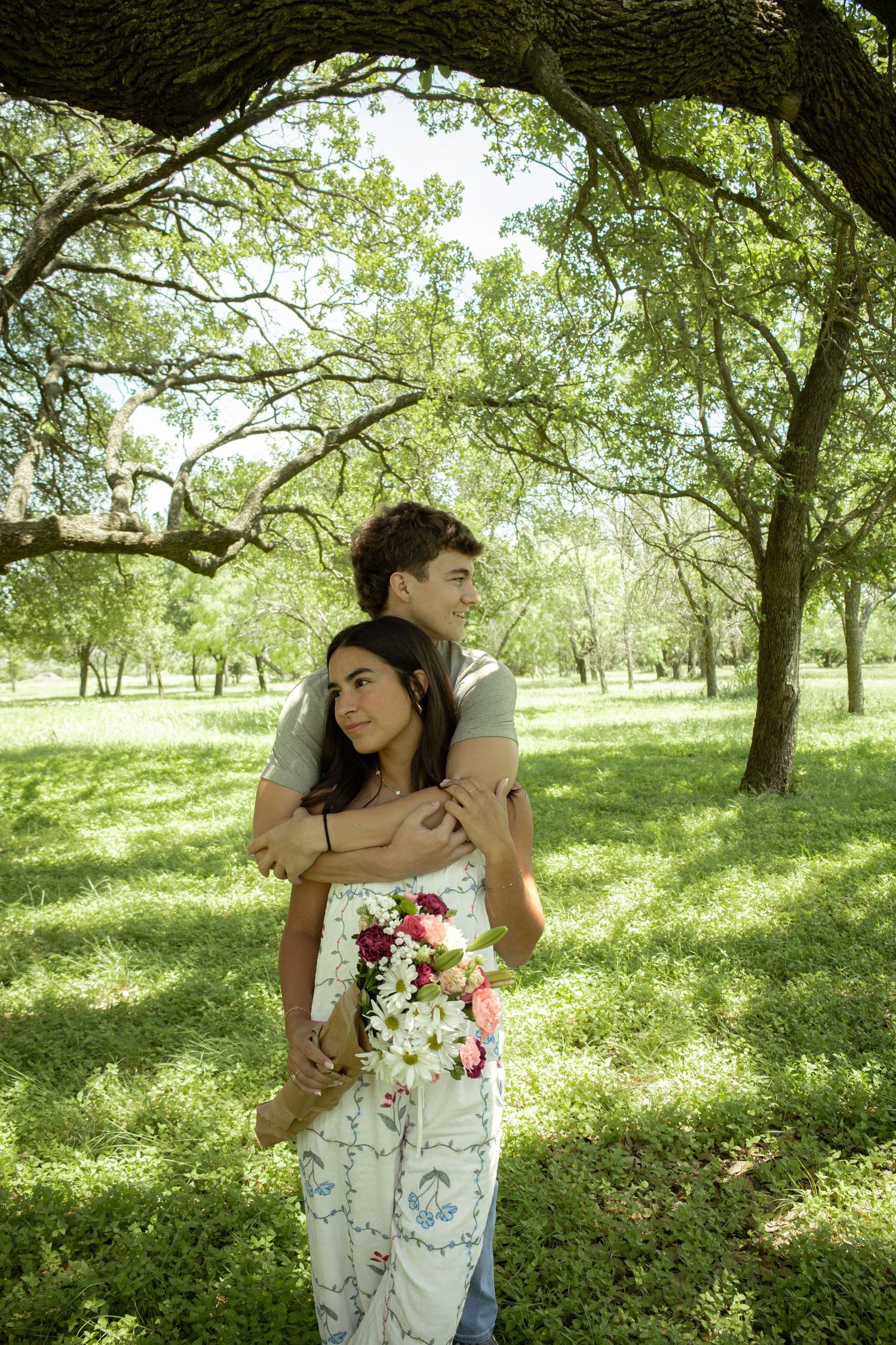 A young couple embraces under oak trees in a green park, the woman holding a bouquet of colorful flowers.
