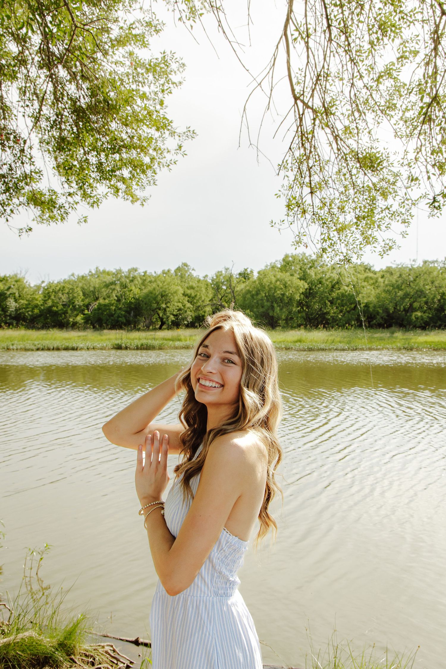 Smiling young woman in a blue dress posing by a serene river surrounded by lush green trees on a bright day.