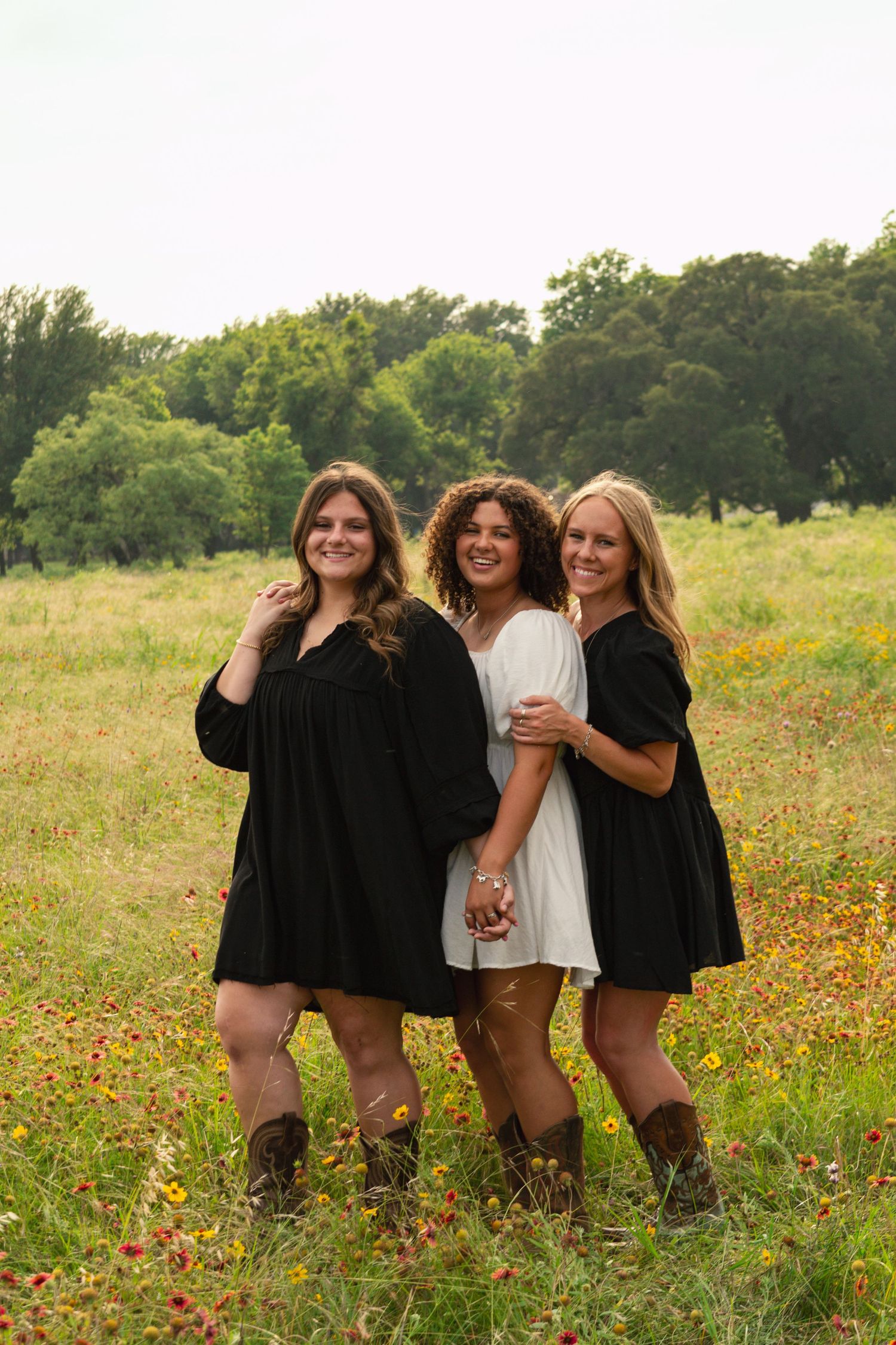 Three young women in black and white dresses posing in a wildflower meadow with cowboy boots.