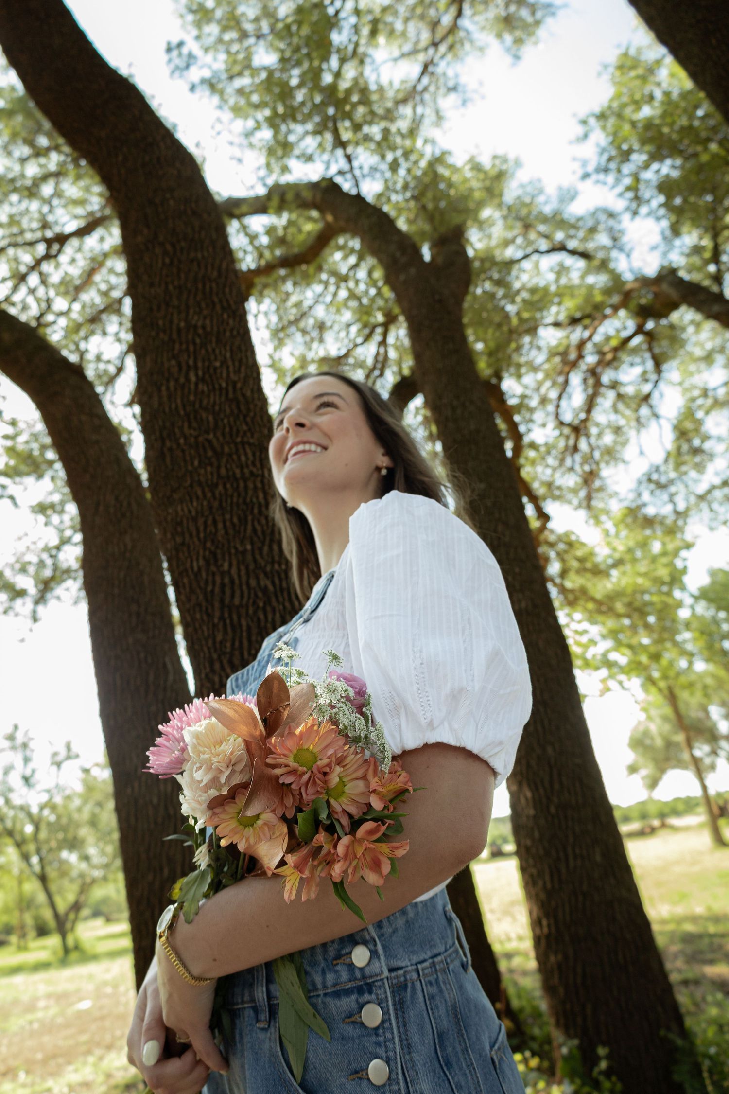 Smiling woman holding a bouquet of colorful flowers outdoors, standing among tall oak trees in a sunny park.