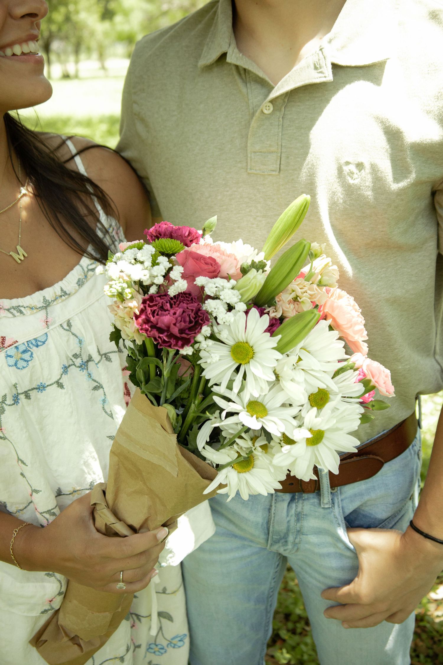 Couple holding a colorful bouquet of daisies, lilies, and pink flowers wrapped in brown paper outdoors.