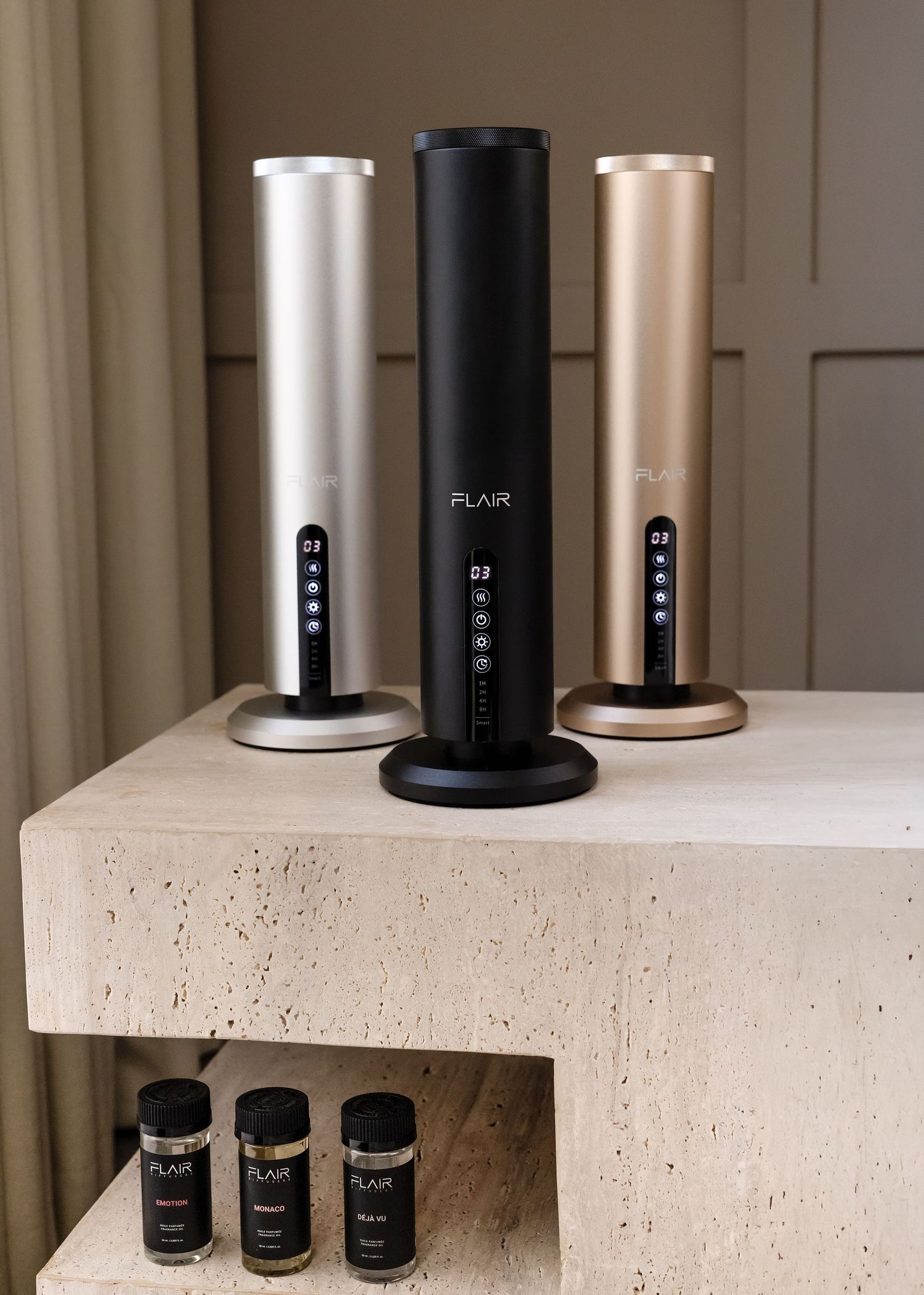 Three Flair brand aroma diffusers in black, silver, and gold displayed on a stone shelf with scent bottles.