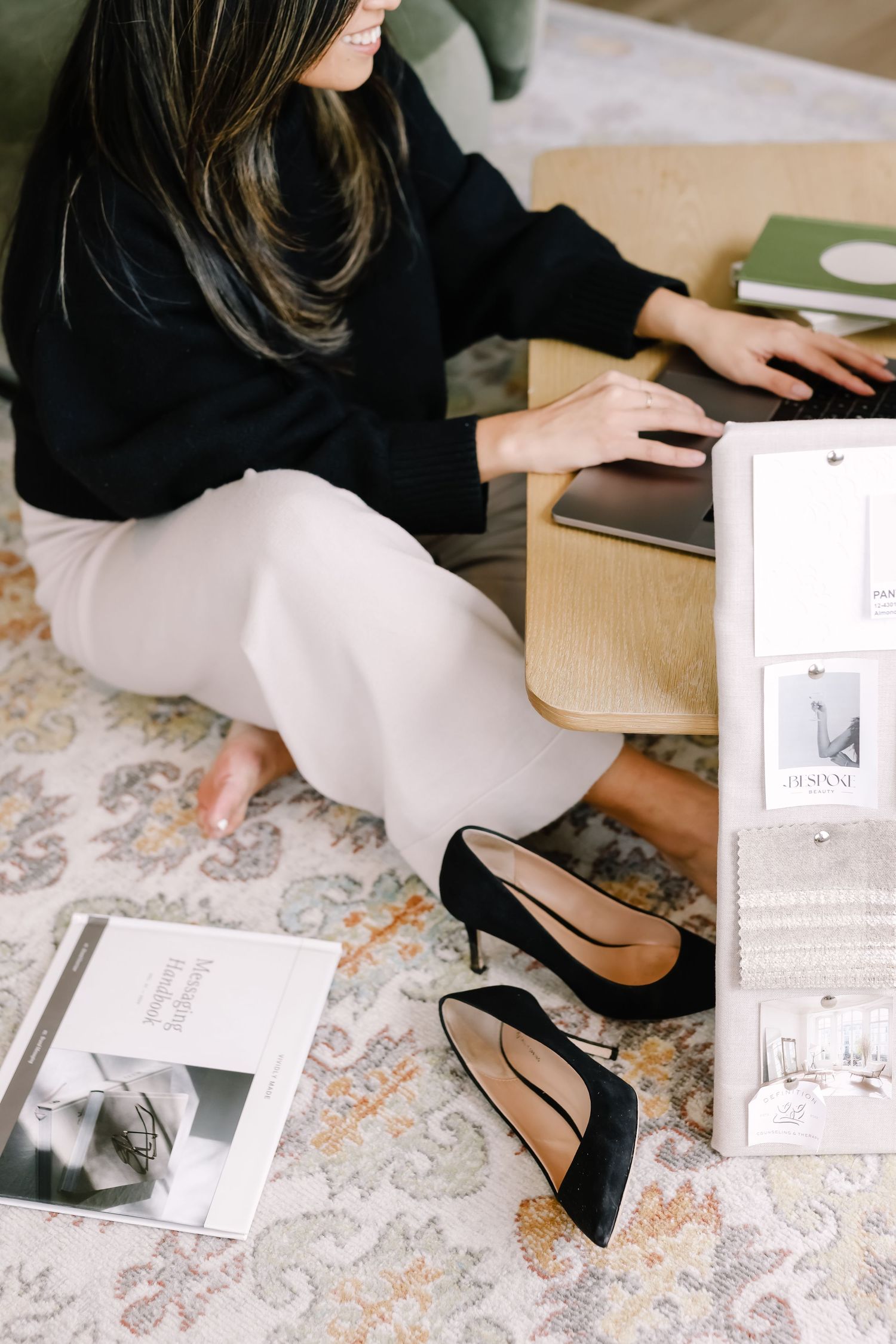 A woman in a black sweater and cream skirt sits on a patterned rug with black heels and design materials beside her.