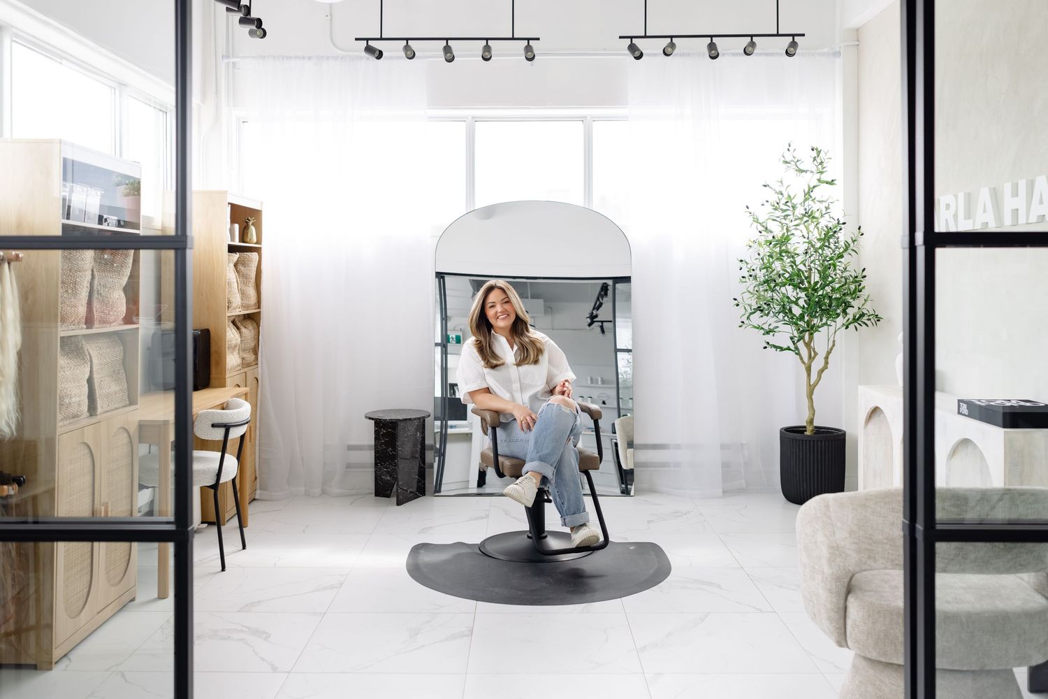 A bright modern home office setup with a grey ergonomic chair on a round mat, indoor plant and wooden shelving unit.
