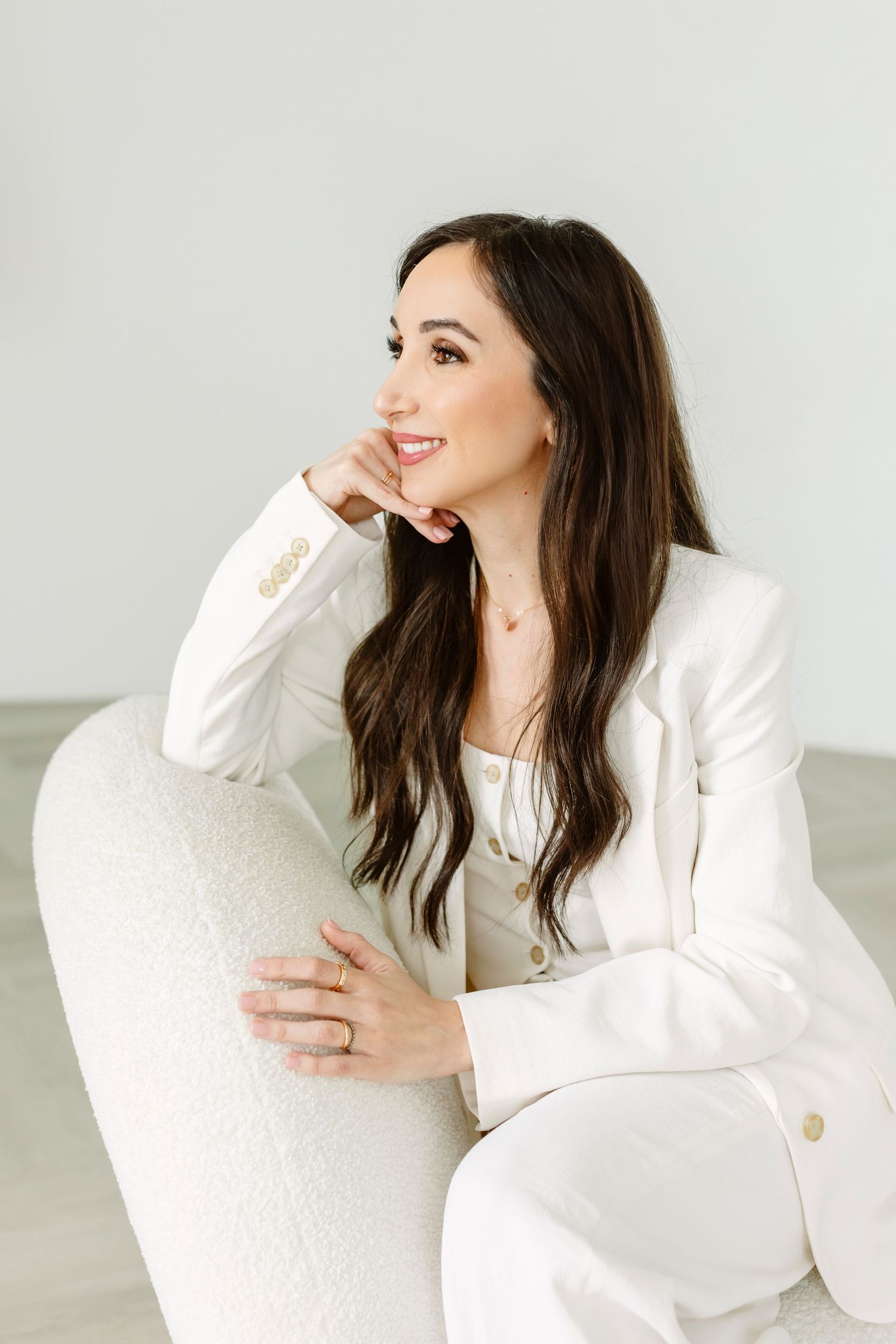 A smiling brunette woman in a white blazer and pants sits elegantly in a white chair against a light background.