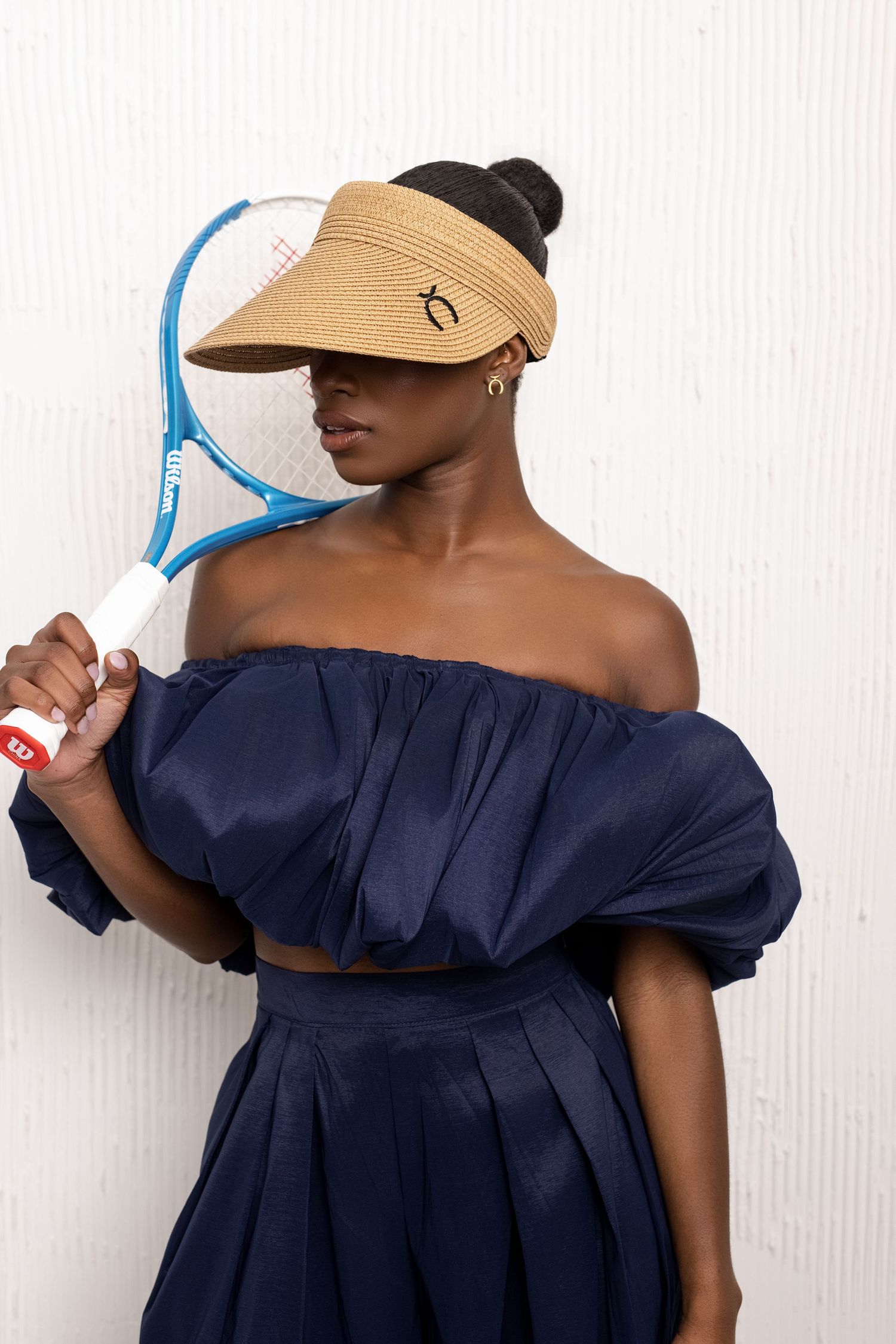Woman in navy off-shoulder outfit holding a blue tennis racket, wearing a straw visor against a white background.