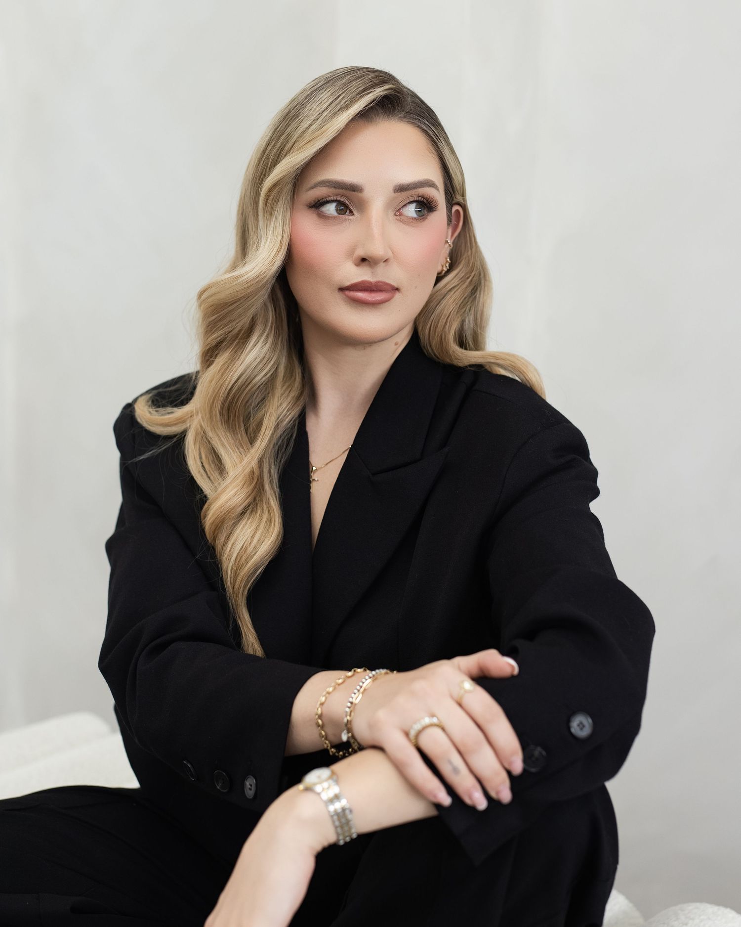 A young woman in a black blazer with blonde wavy hair and gold jewelry poses confidently against a light background.