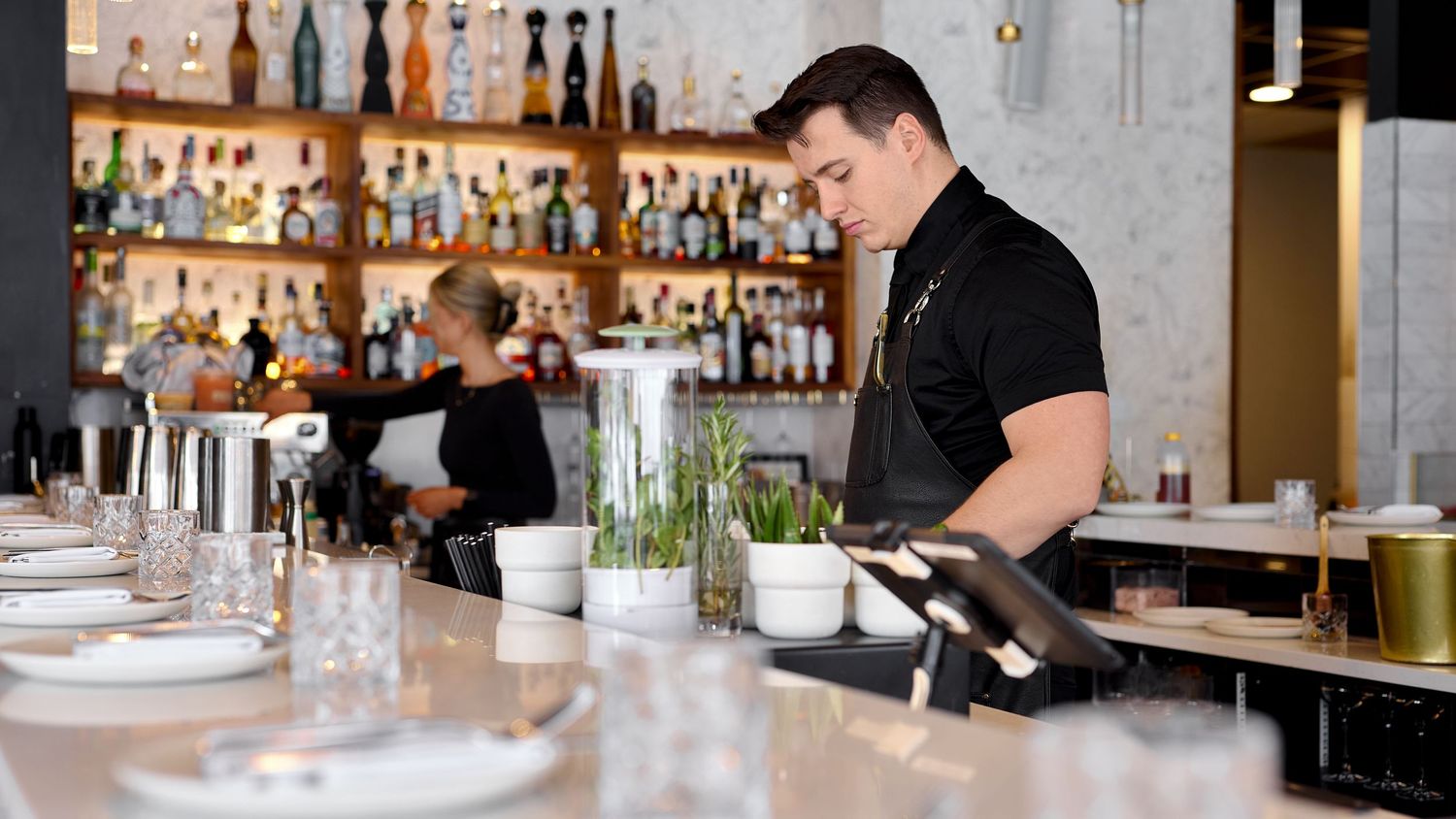 Bartender in black uniform preparing drinks at an elegant restaurant bar with glassware and bottle display.