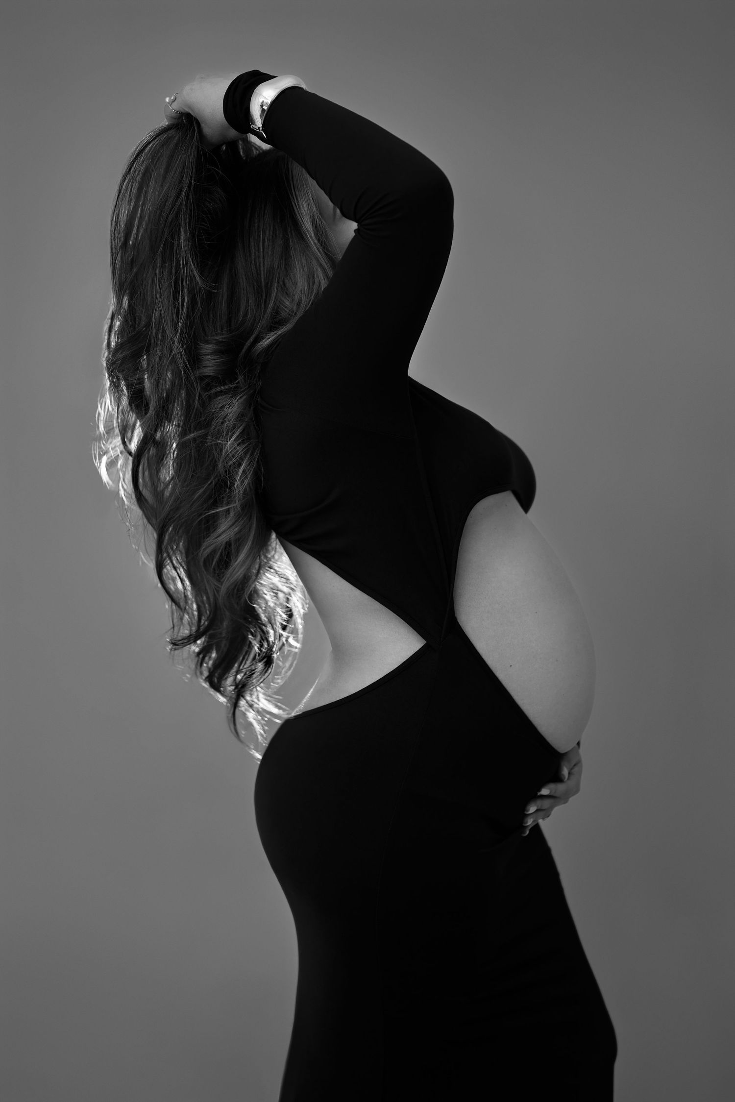 Artistic black and white photo showing silhouette of figure wearing black dress with cutout back design against grey background.