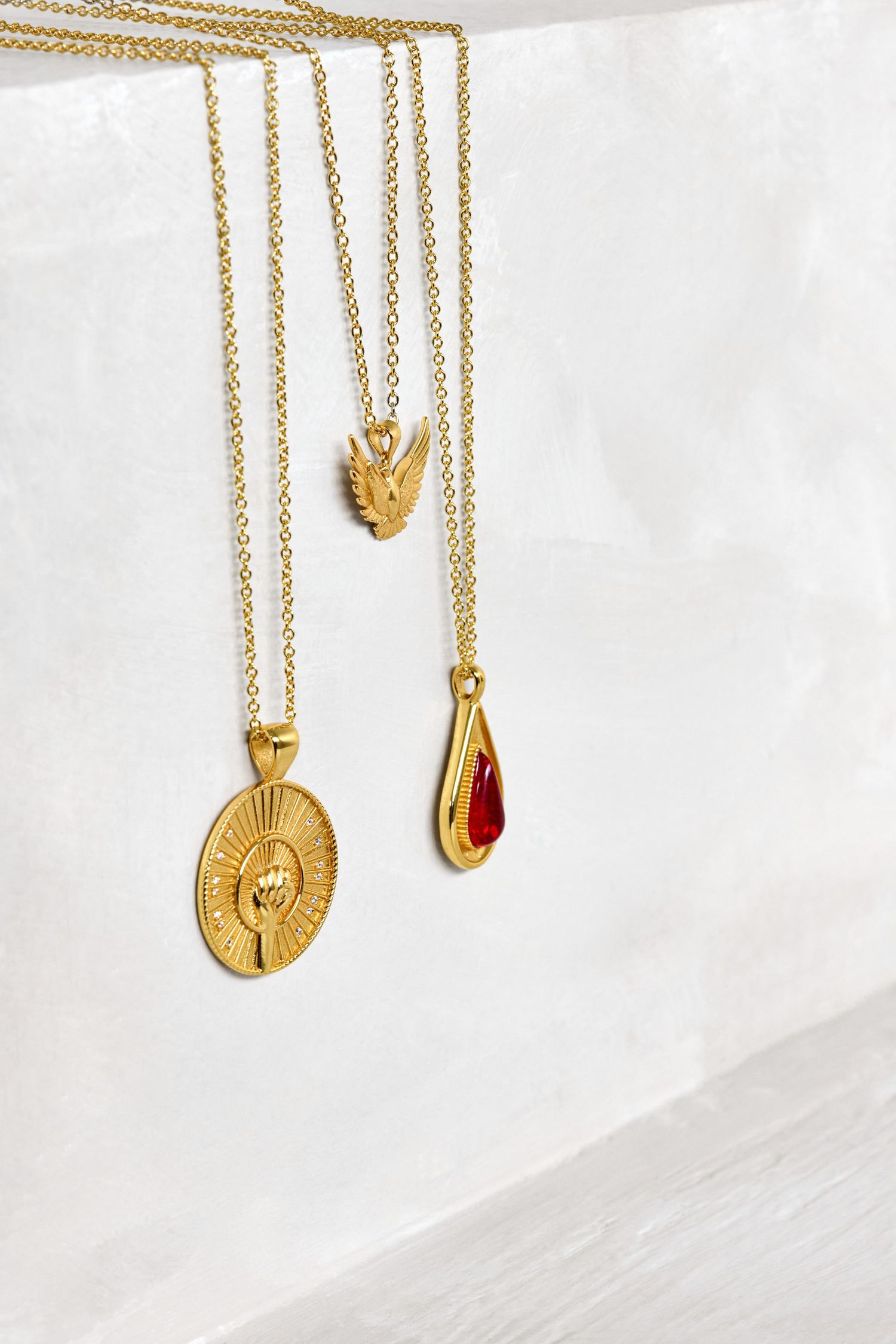 Three gold pendant necklaces on white background featuring teardrop, medallion and wing charms.