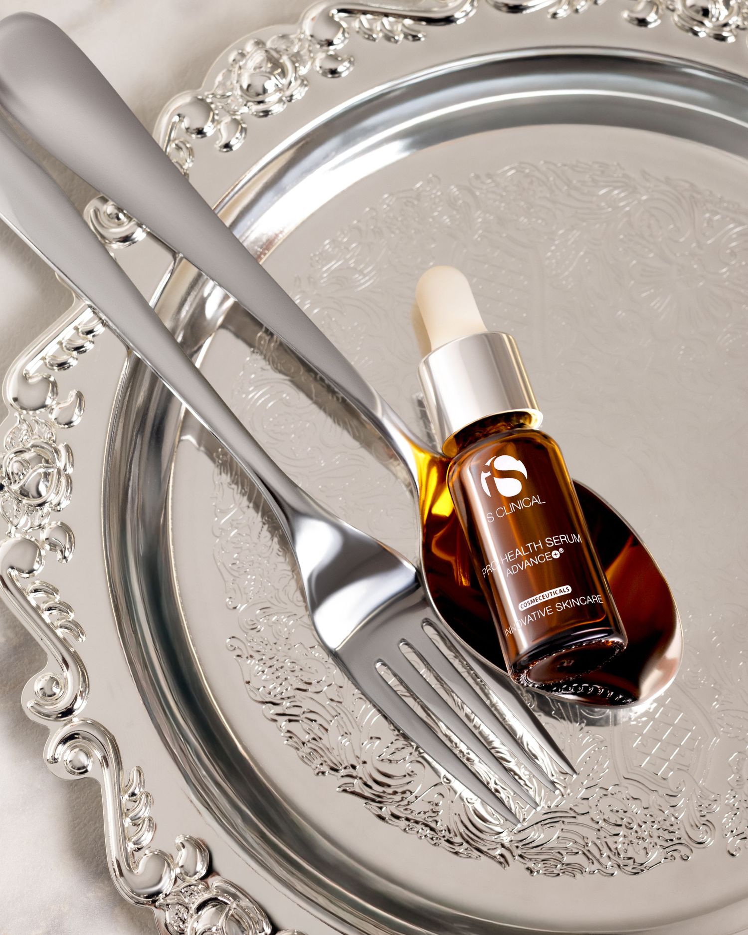 iS Clinical Youth Serum dropper bottle on a silver decorative tray with elegant silverware fork and spoon.