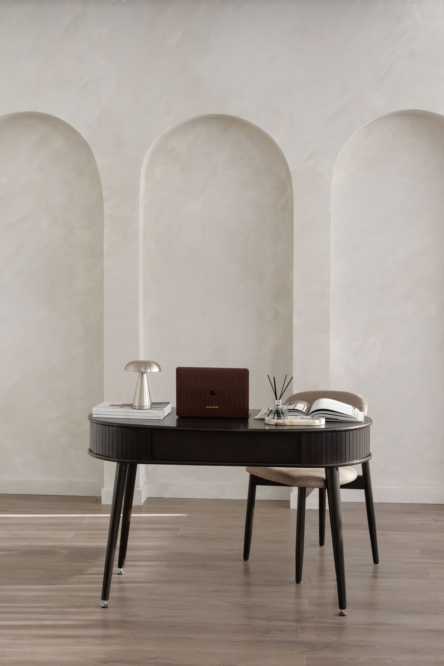 Minimalist home office with black oval desk, brown laptop, table lamp, and beige chair against arched wall niches.