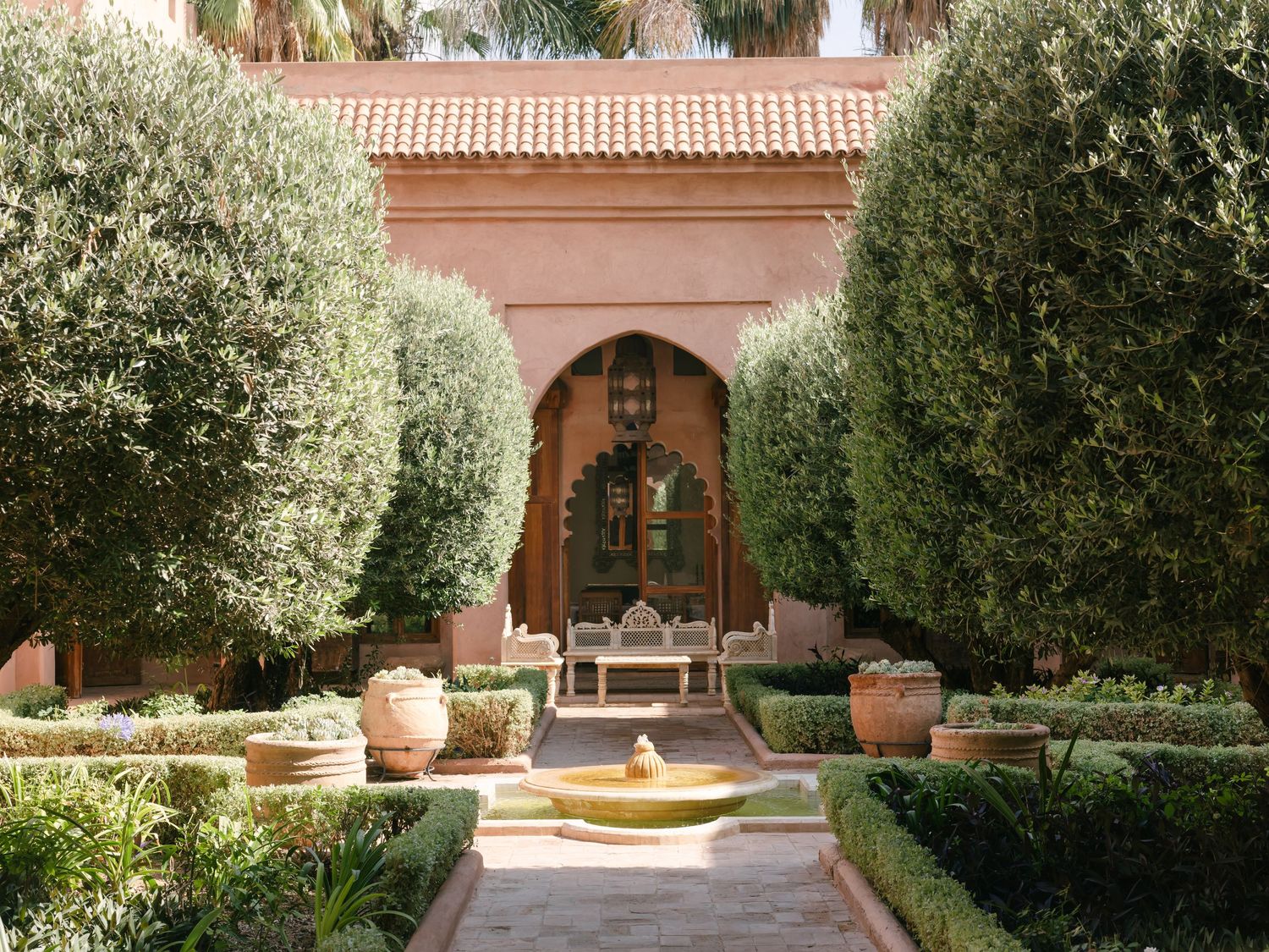 Lush Moroccan courtyard garden with trimmed trees, terracotta pots, fountain, and ornate arched doorway in warm pink tones.