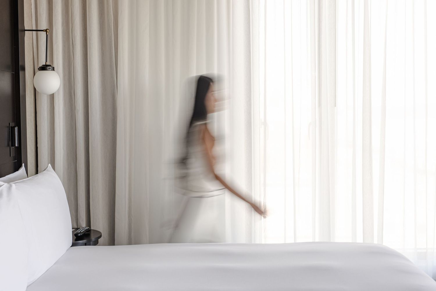 Blurred motion of a bride in a white dress and veil moving near sheer curtains in a bright hotel room.