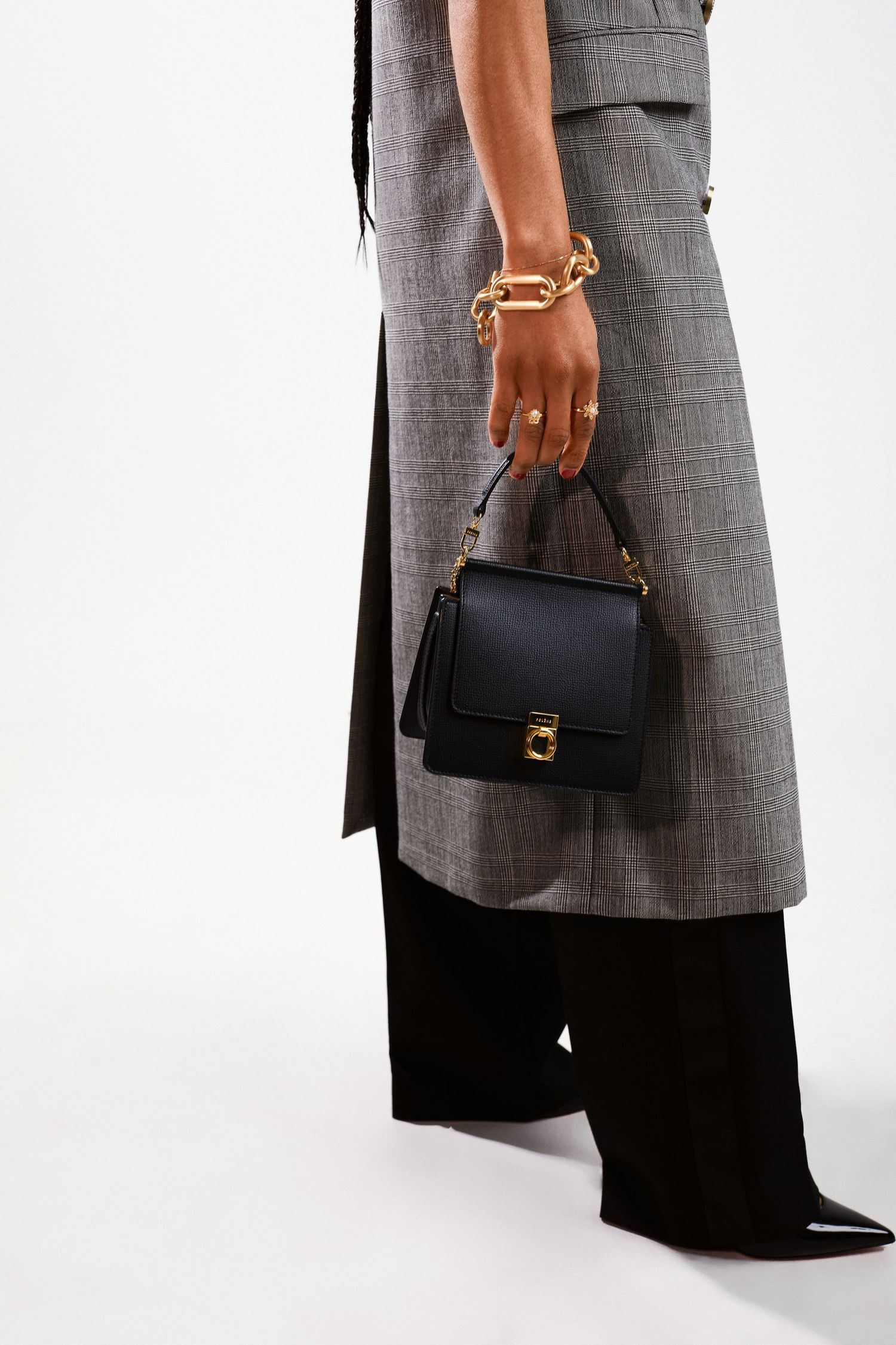 Model holding a small black leather top-handle bag with gold hardware, wearing a gray plaid coat and black wide-leg trousers.