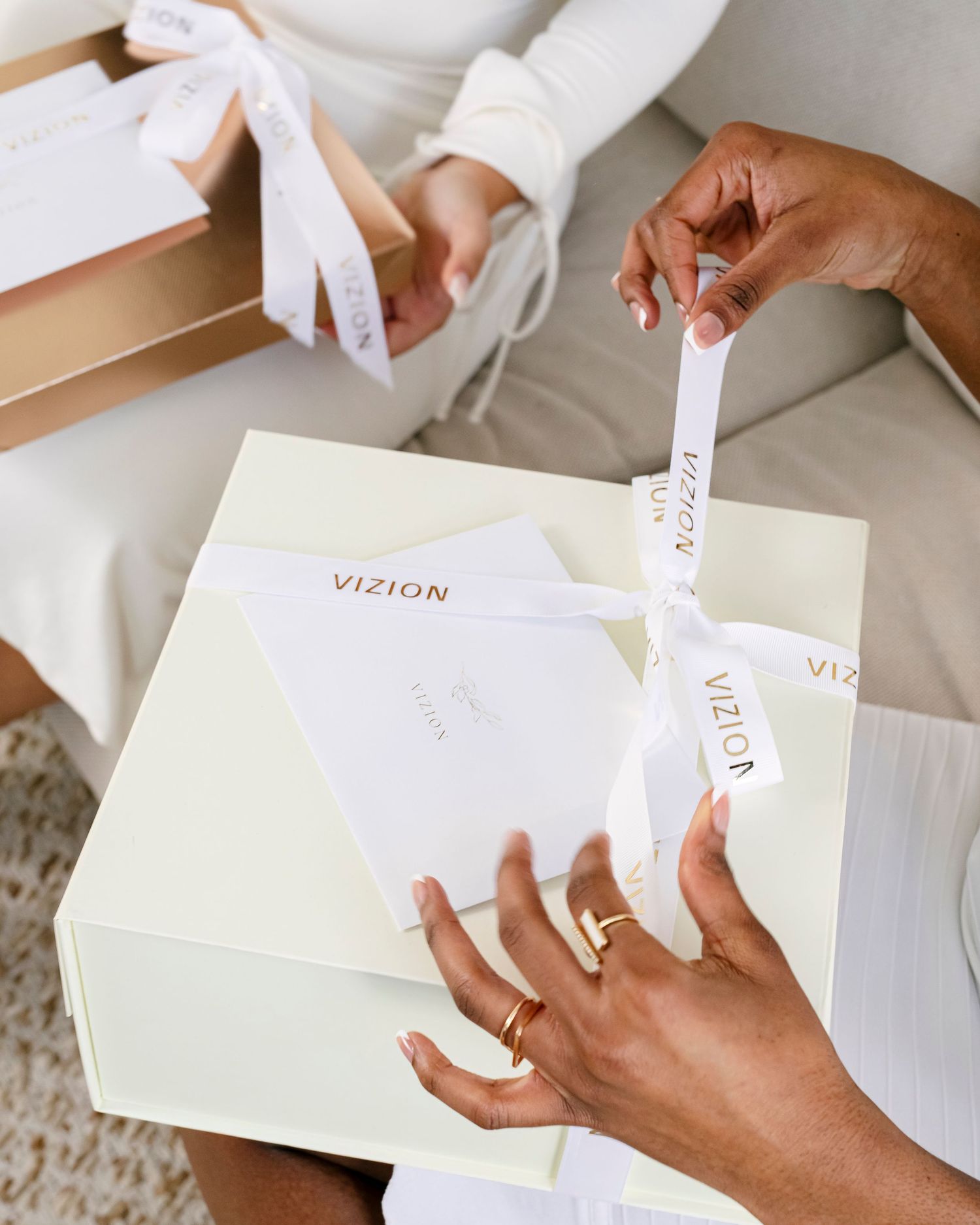 Two people opening elegant cream and gold VIZION gift boxes tied with white satin ribbons.