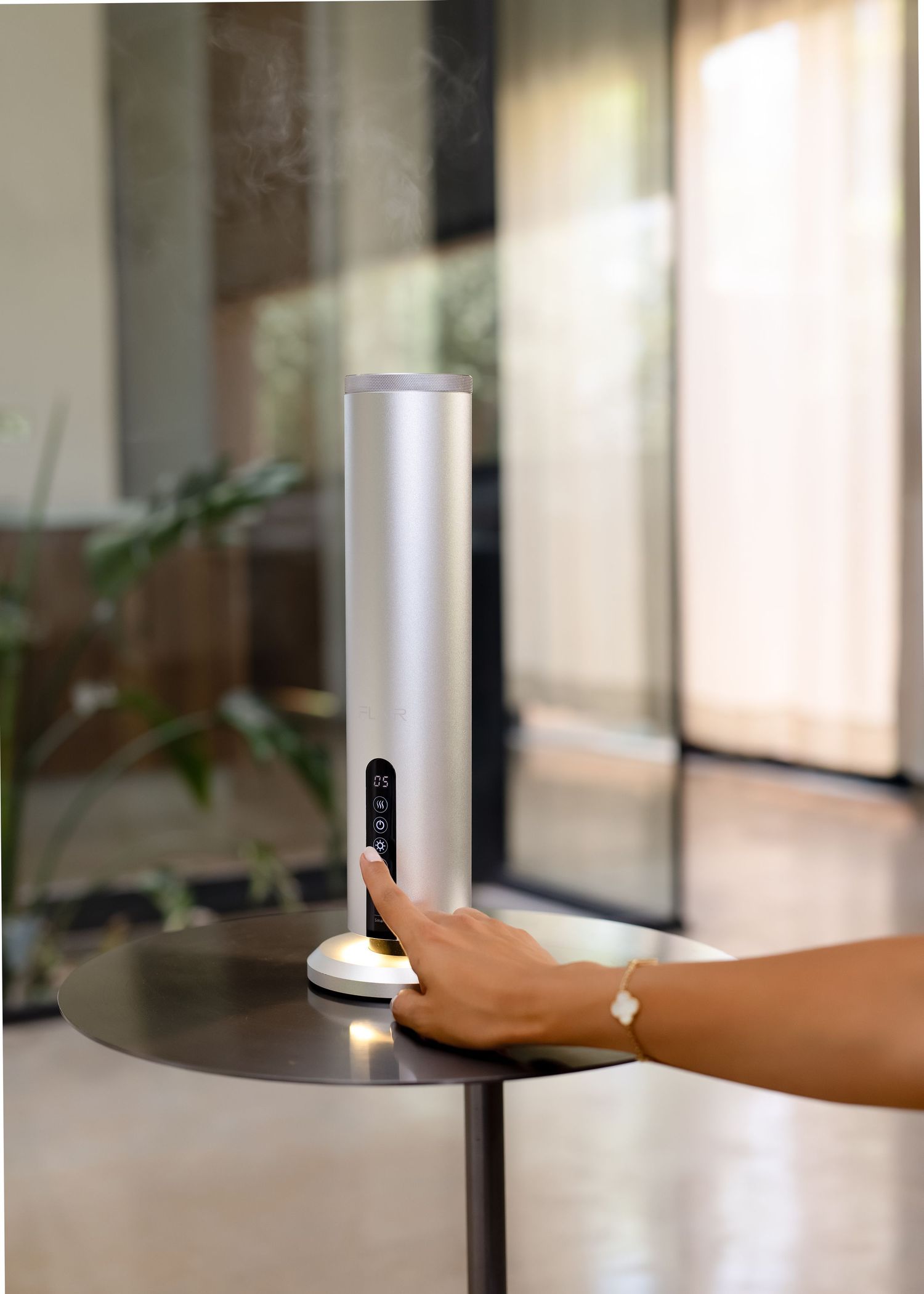 Hand touching a sleek silver cylindrical aroma diffuser on a modern side table in a bright interior setting.