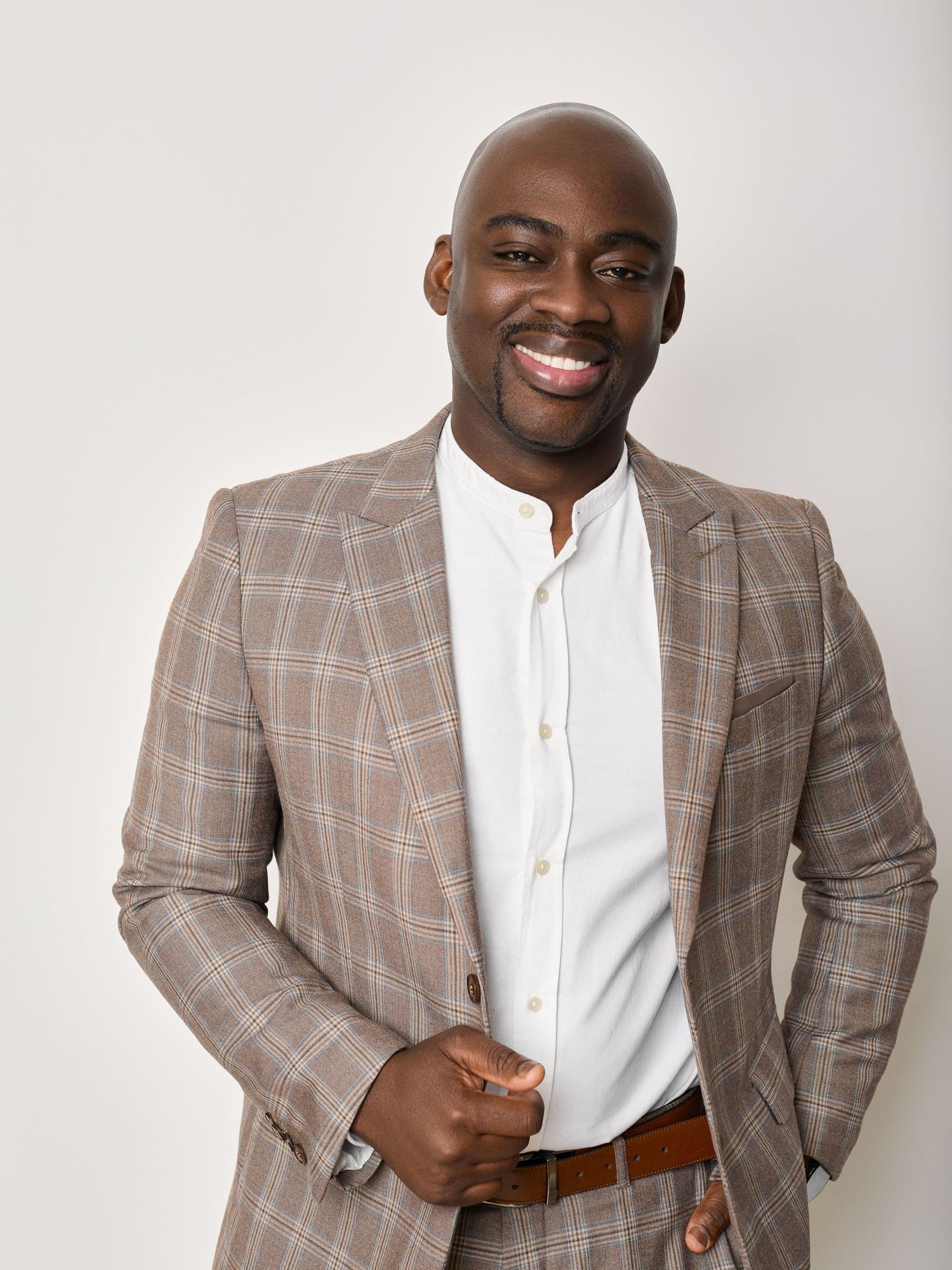 A person in a plaid brown blazer and white shirt poses confidently against a light background in a professional photoshoot.