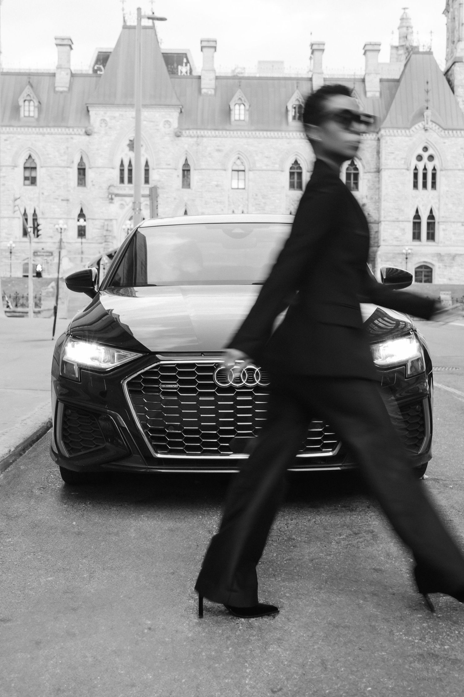 A black and white photo sequence shows a blurred figure walking past a parked Audi car in front of a castle-like building.