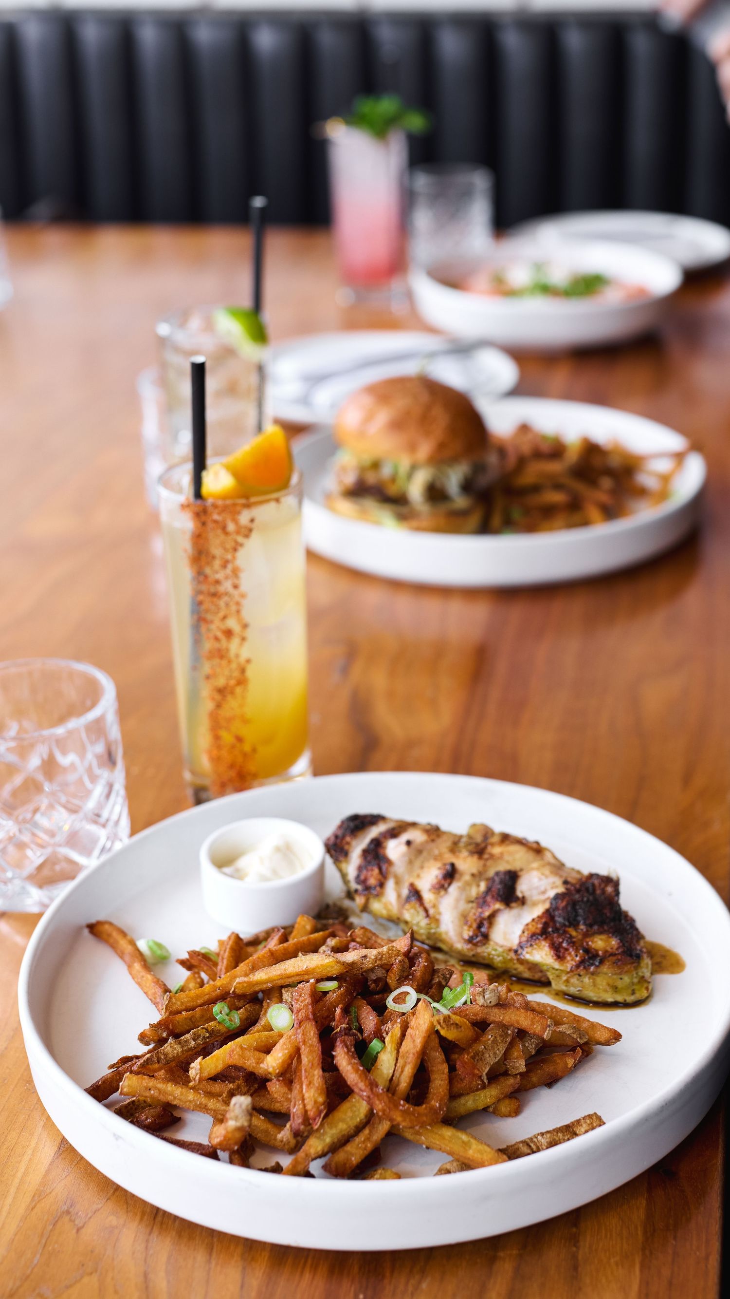 Restaurant table with grilled chicken, seasoned fries, a burger, and a citrus cocktail served on white plates.