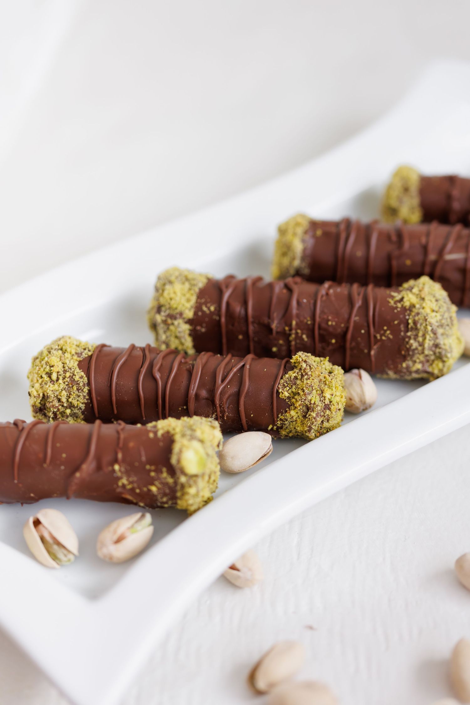 Chocolate covered pistachio cannoli with crushed nuts sprinkled on the ends displayed on a white surface.