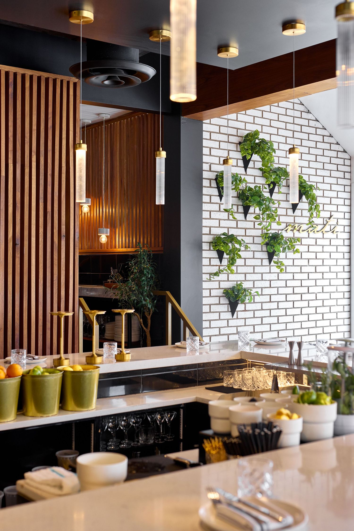 Modern restaurant interior with white brick walls, wooden slat dividers, pendant lights and green plants on marble counters.