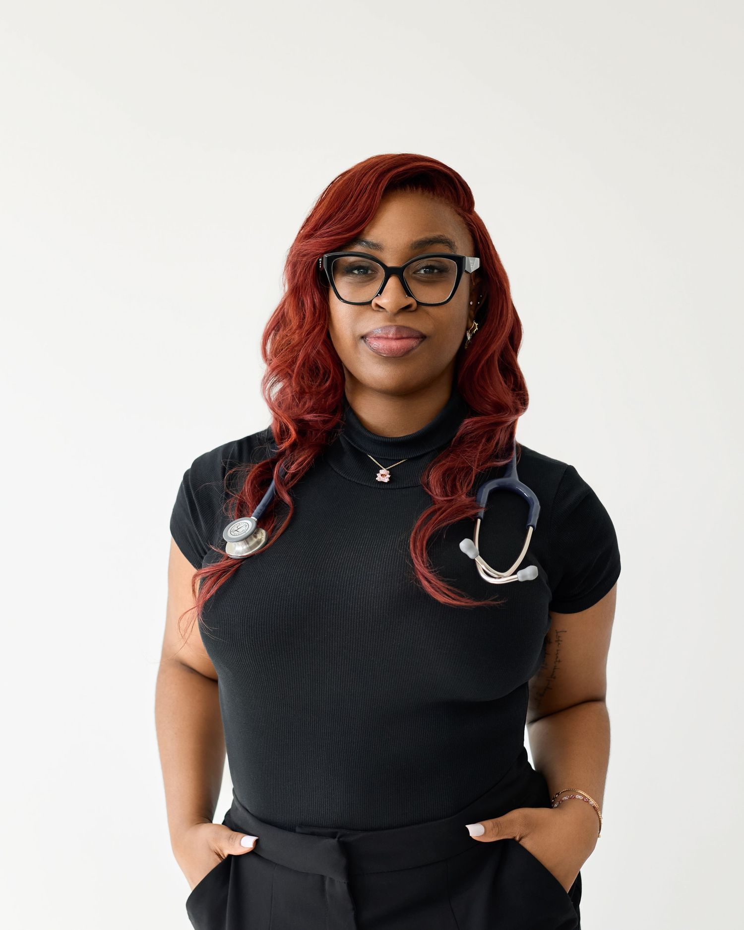 Healthcare professional with red hair and glasses wearing black scrubs poses confidently against white background.