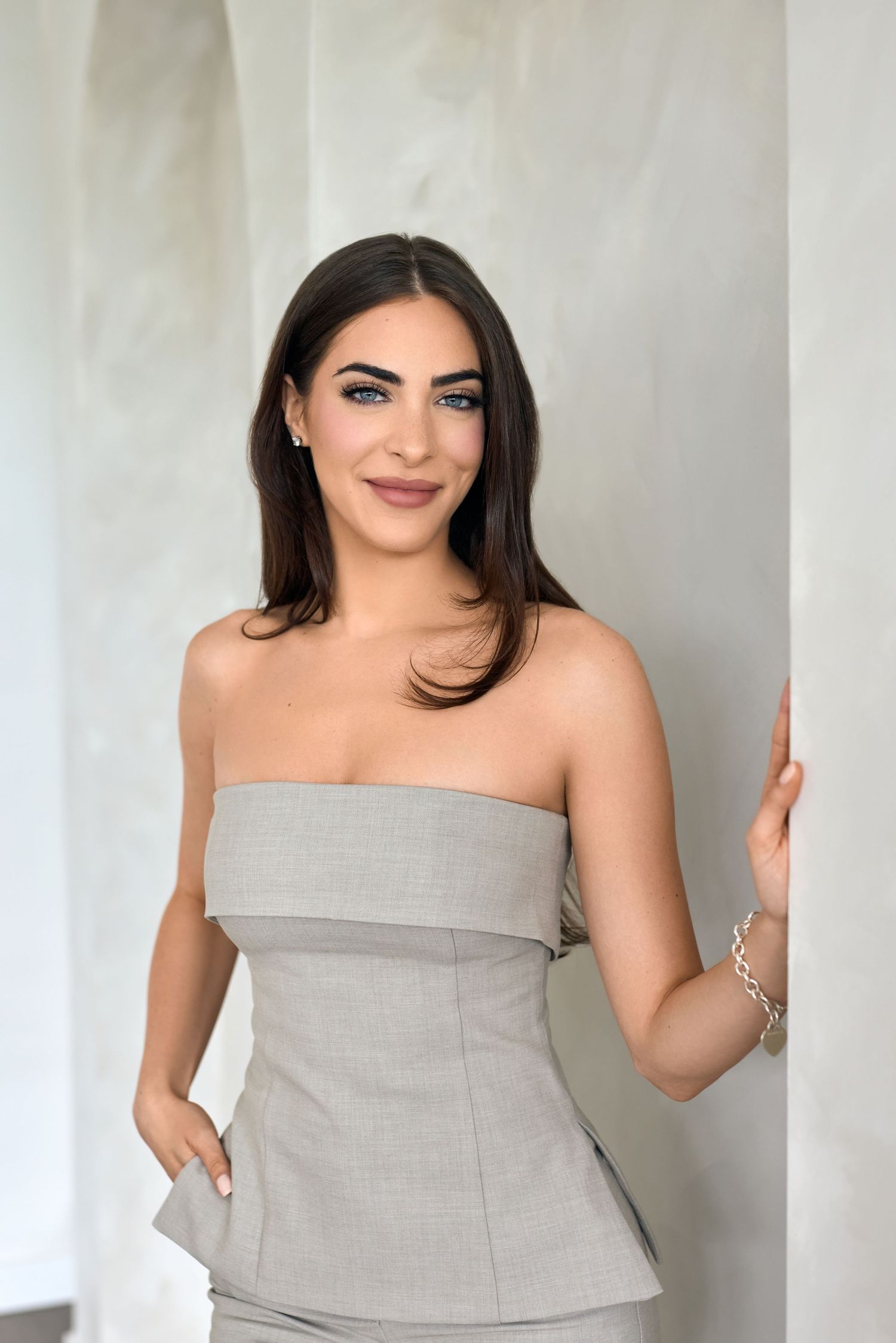 Woman in strapless grey structured top poses elegantly against white wall.