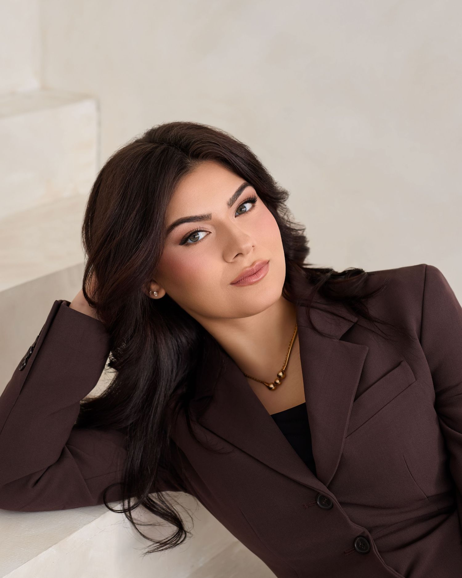 A young woman in a brown blazer with dark hair poses elegantly, wearing a gold necklace against a white background.