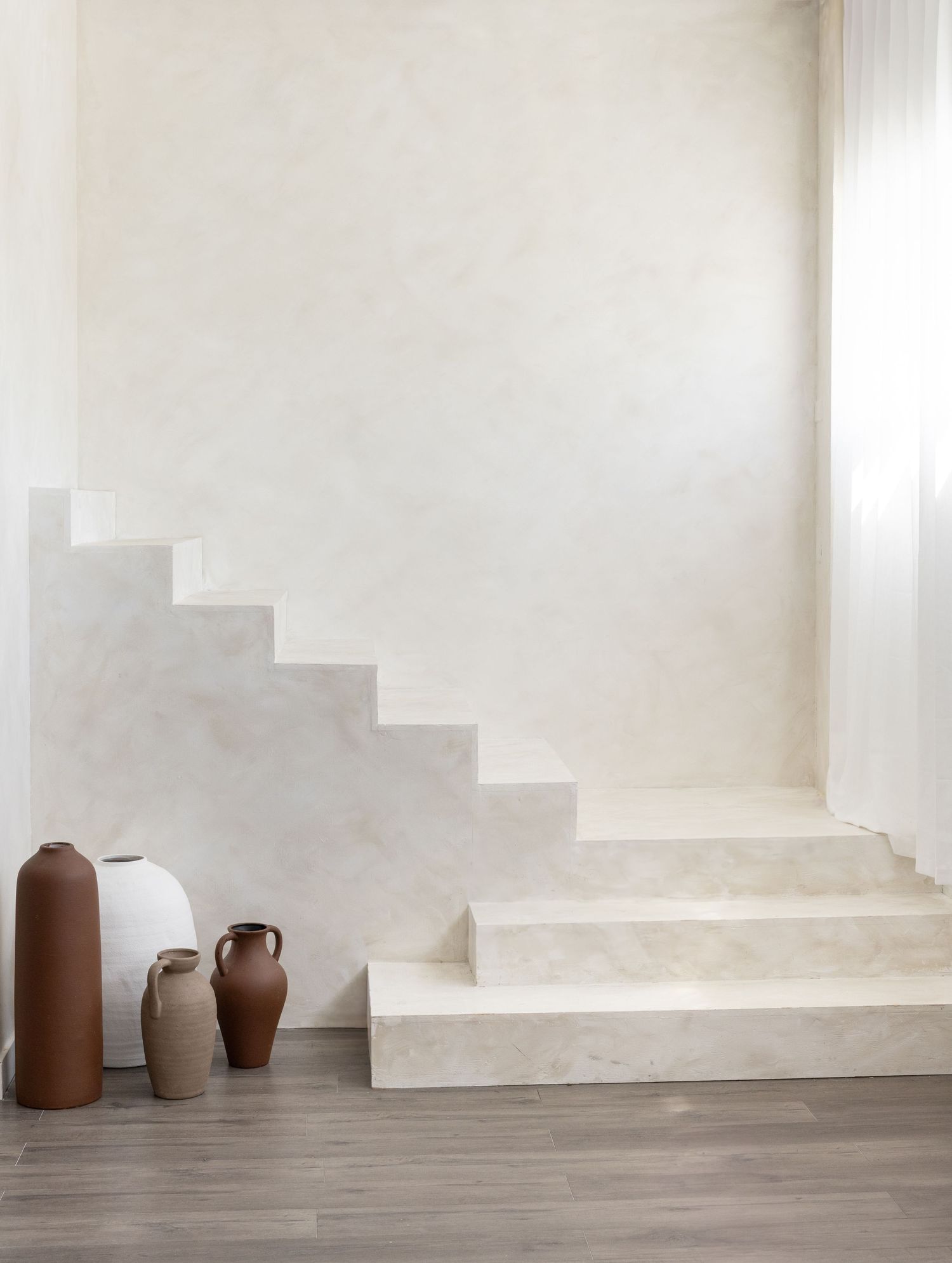Minimalist white stone staircase with warm terracotta ceramic vases on wood floor in a serene, neutral-toned interior space.