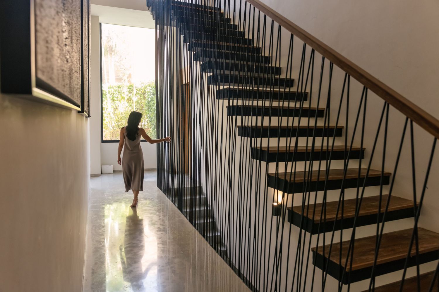 Woman in flowing dress walking near modern floating staircase with black metal railings in bright contemporary home.