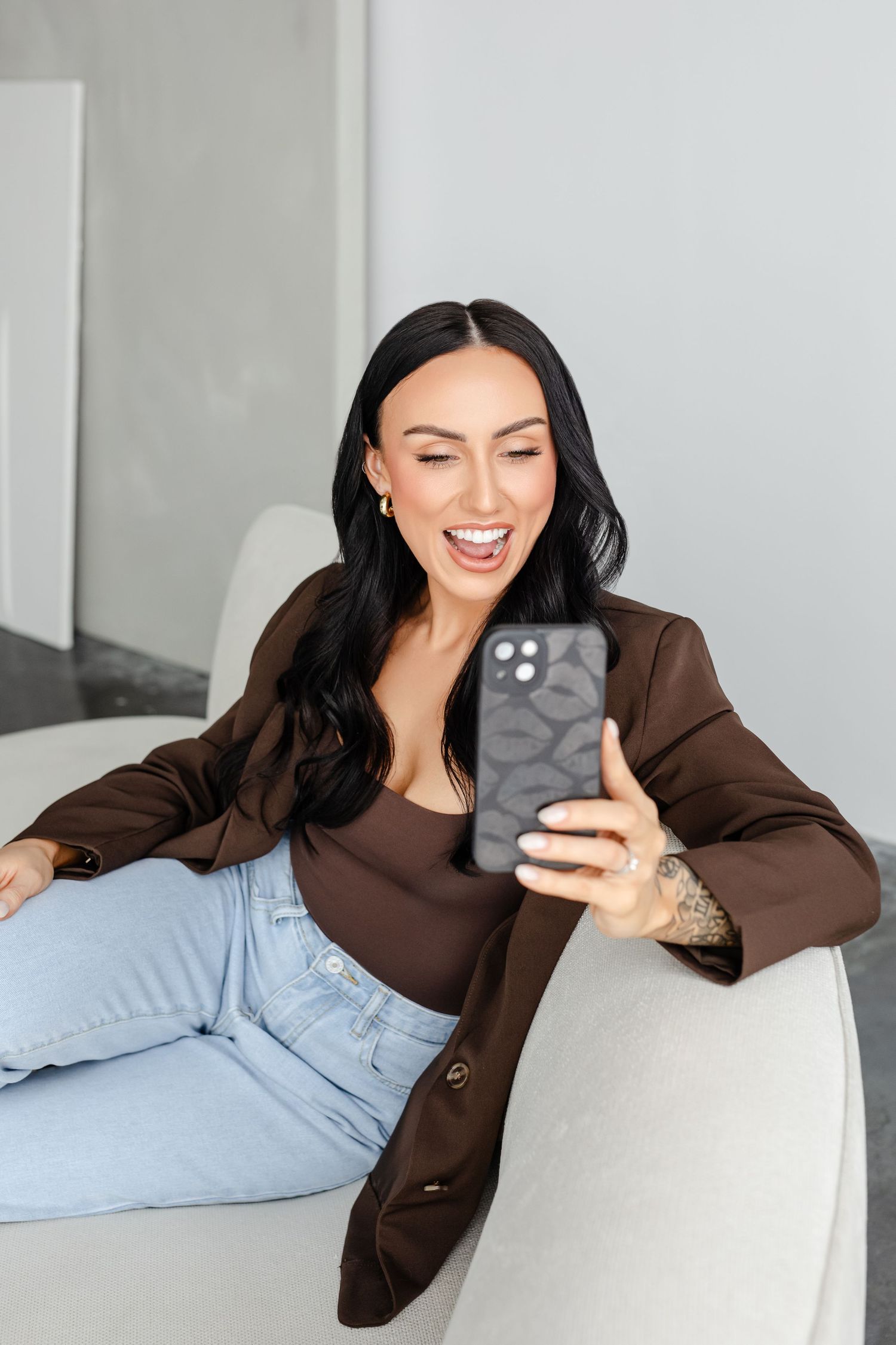 Woman in brown cardigan and light jeans relaxes on white couch while looking at smartphone with animated expression.