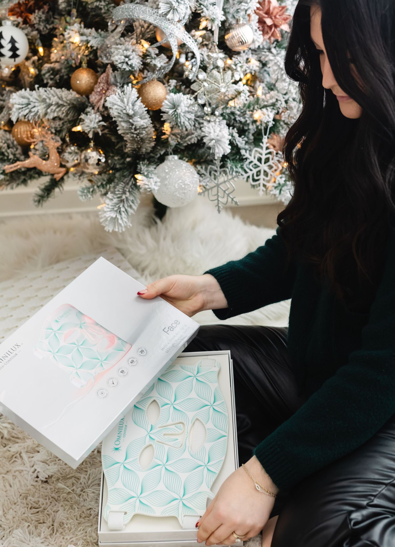 Woman in green top opening a gift box near a decorated Christmas tree with white and rose gold ornaments.