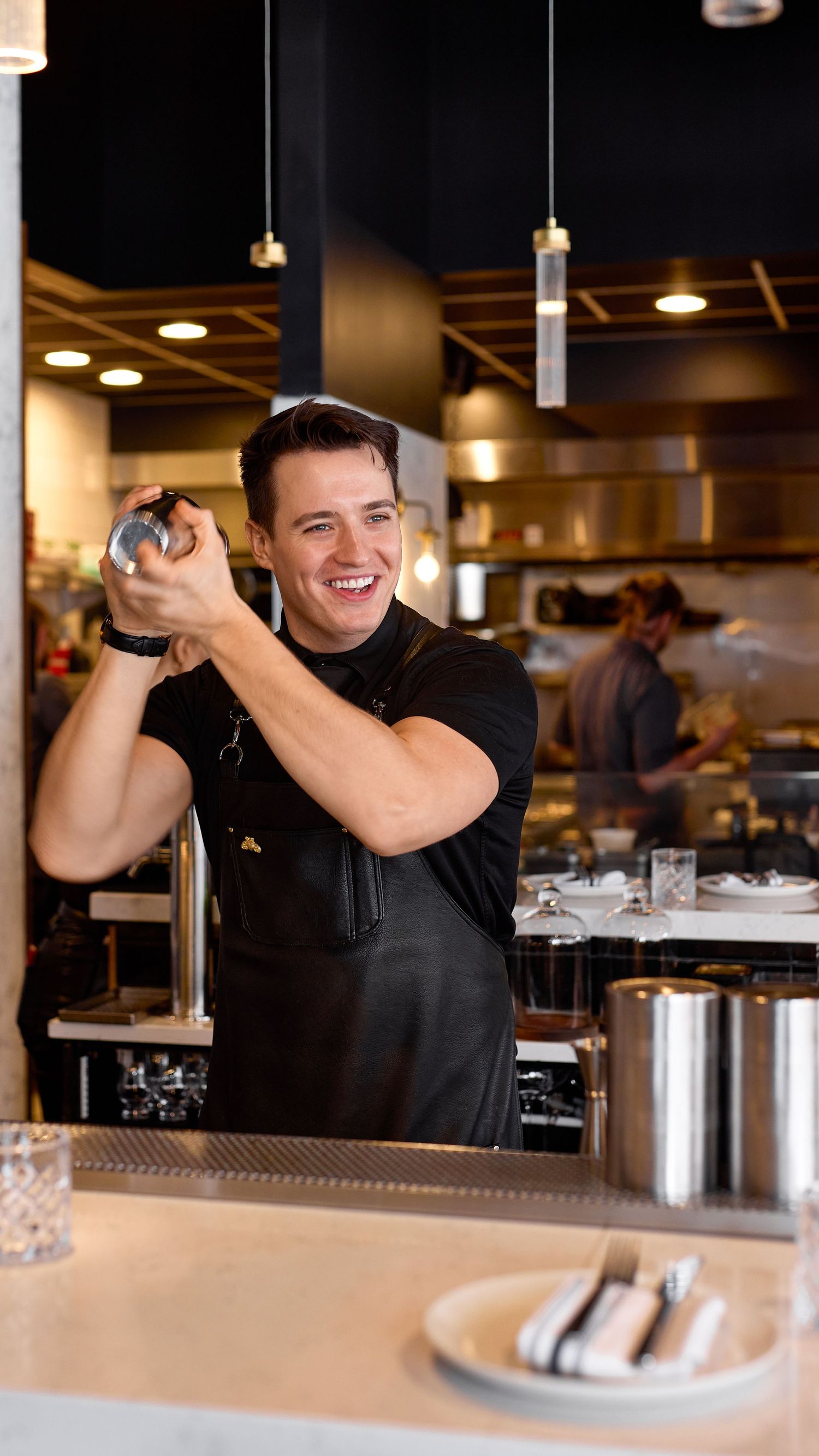Smiling bartender in black apron shaking a cocktail mixer in a modern restaurant kitchen setting.