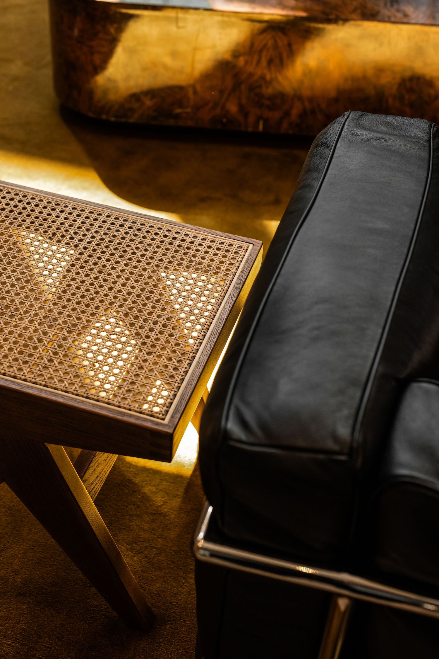Elegant interior with black leather chair, rattan side table with gold frame, and burl wood ottoman in warm lighting.