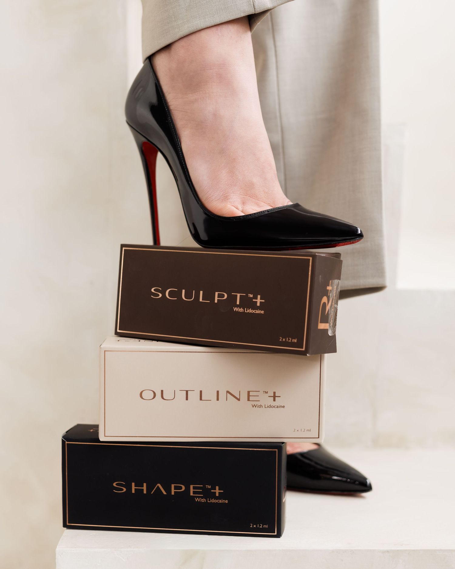 Black patent leather heels stepping on luxury beauty product boxes.