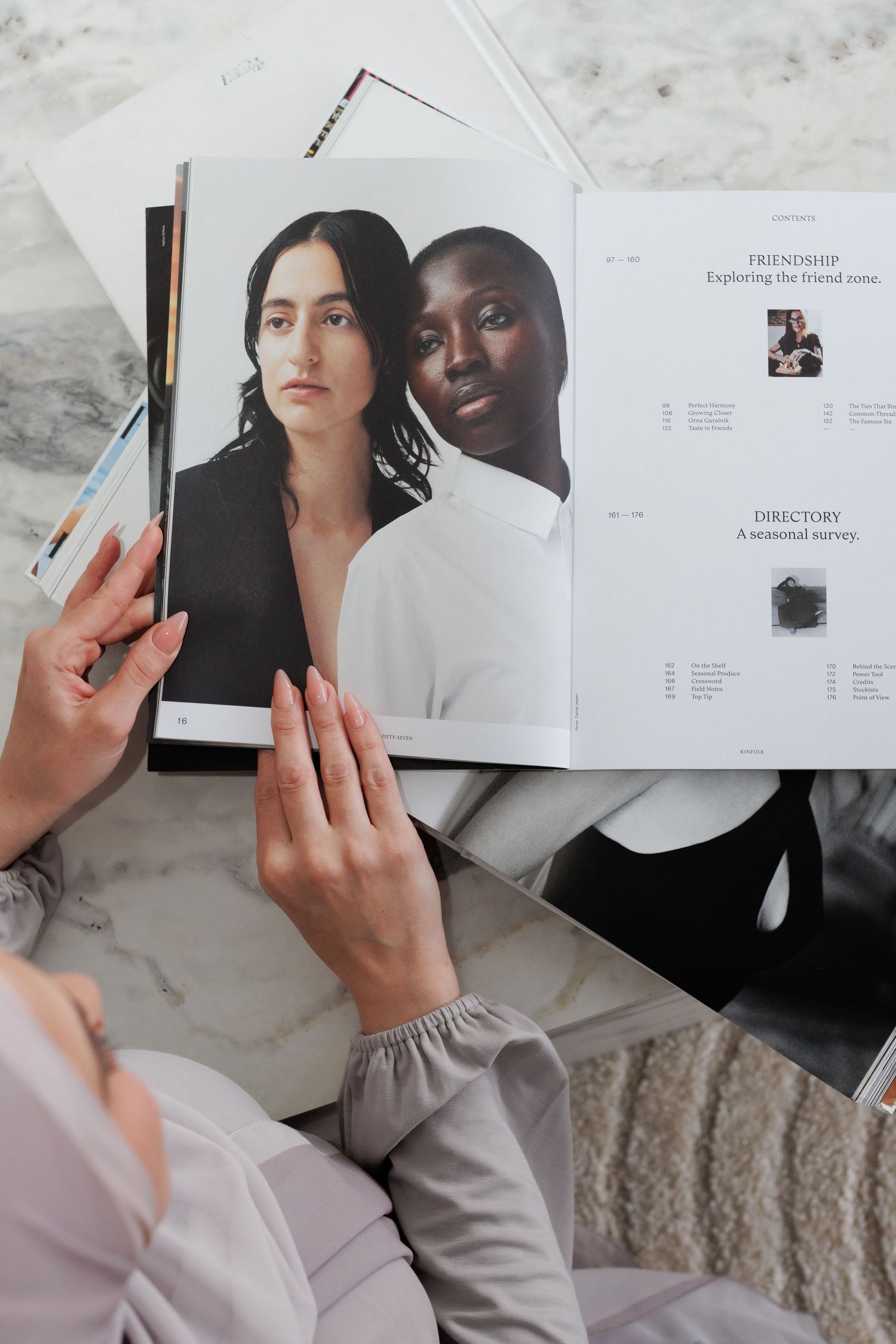 Person reading a fashion magazine featuring two women models, one in black and one in white top, on a marble surface.
