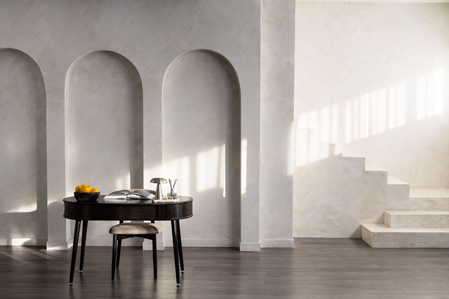 Minimalist room with arched wall niches, dark desk, stool, and marble staircase bathed in natural light.