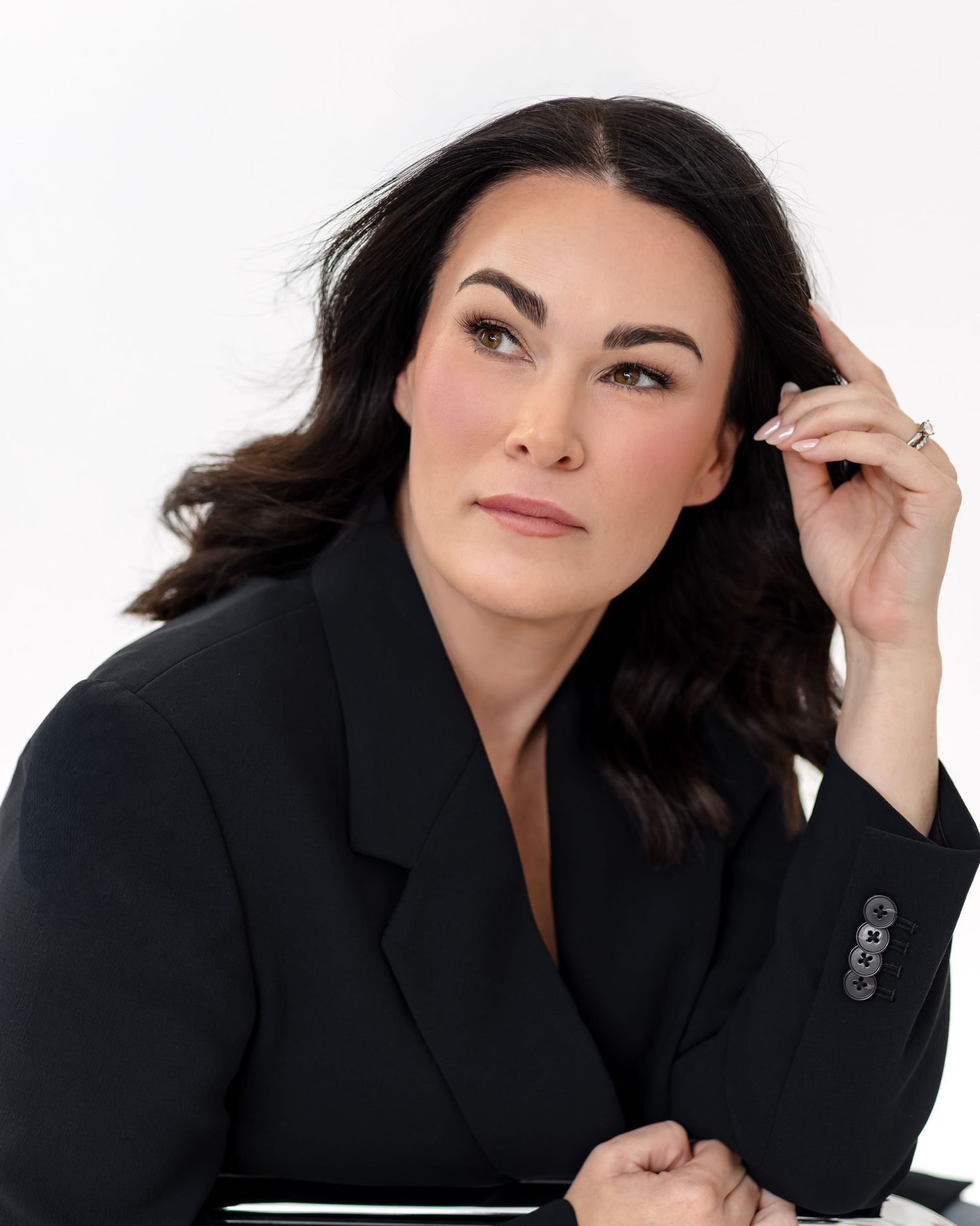 Professional woman in black blazer with dark wavy hair poses confidently against white background.