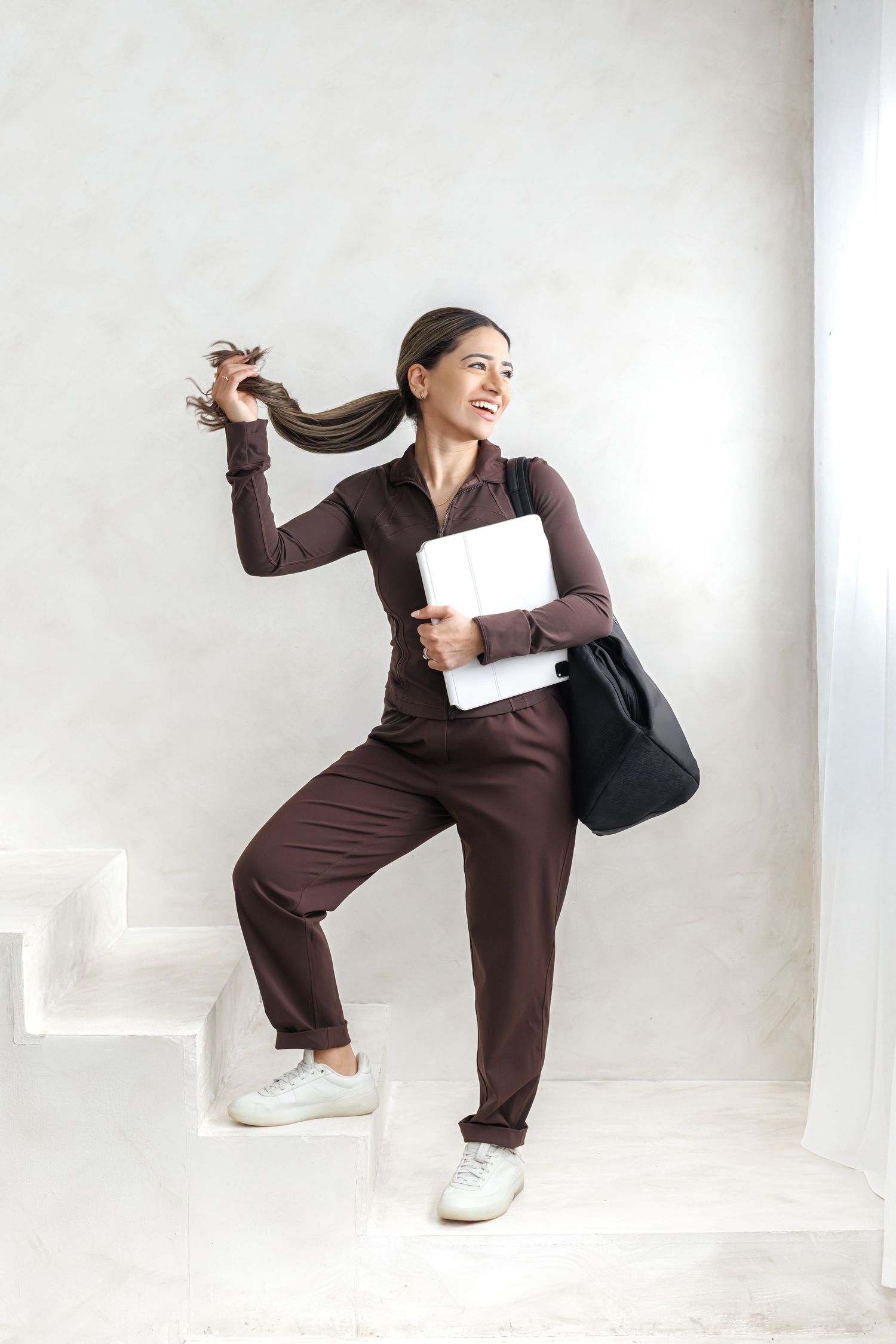 Smiling woman in brown tracksuit climbing stairs, holding laptop and bag, playfully tossing her ponytail.