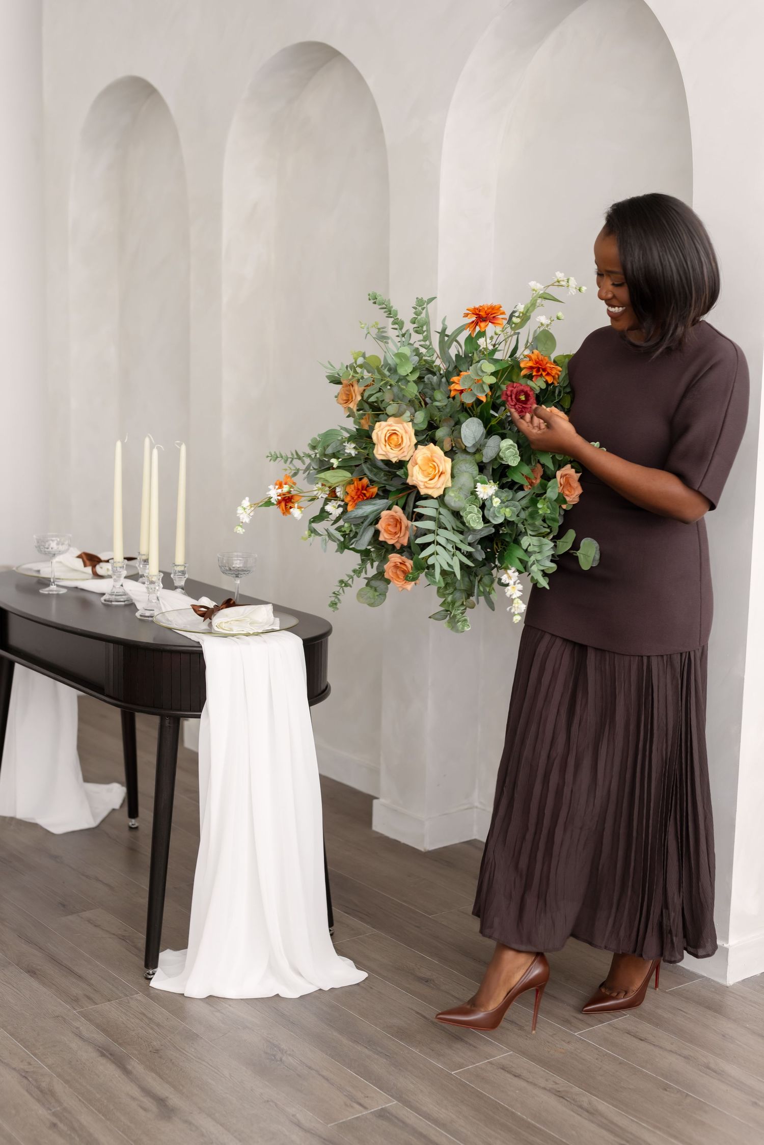 A person in a brown dress arranges a floral centerpiece on a white-draped table in an elegant room with arched walls.