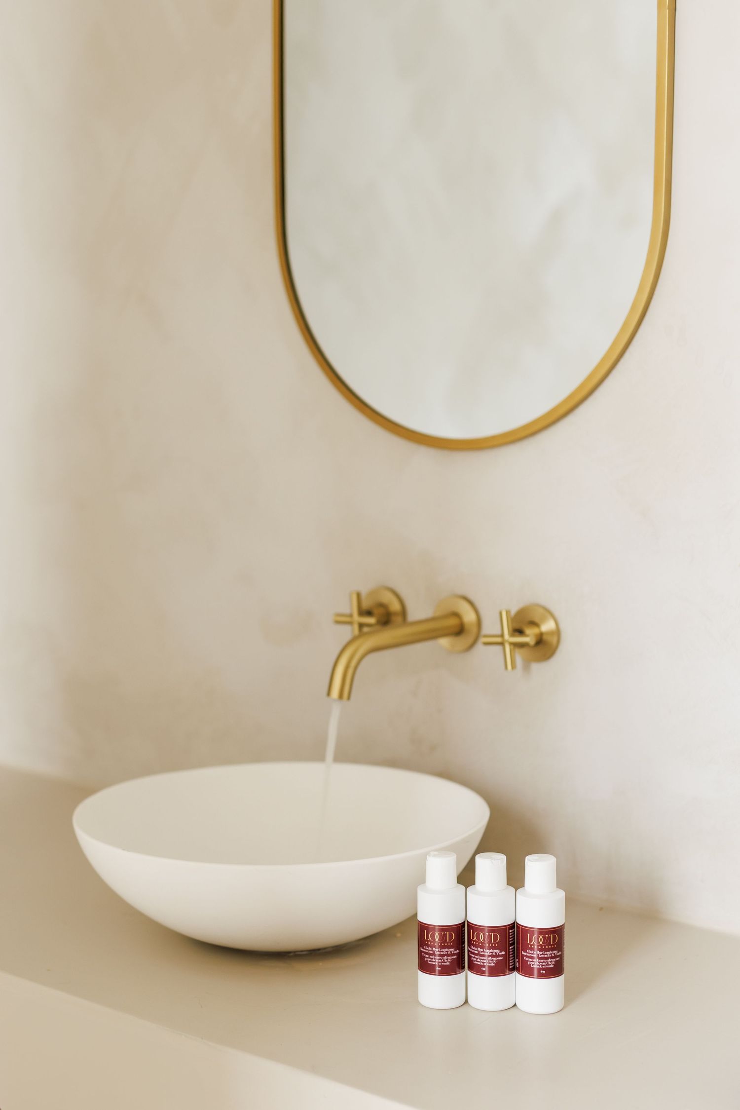 Modern minimalist bathroom with white vessel sink, gold wall-mounted faucet, oval mirror, and three skincare product bottles.