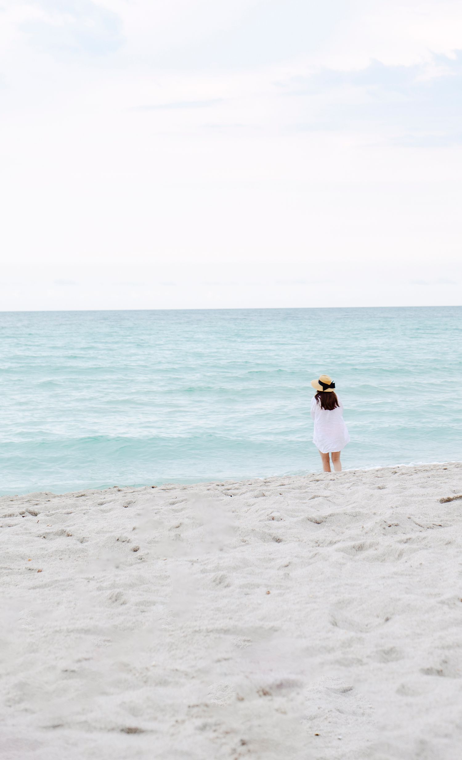 Person in white clothing walks along pristine white sandy beach with turquoise ocean waters in the background.