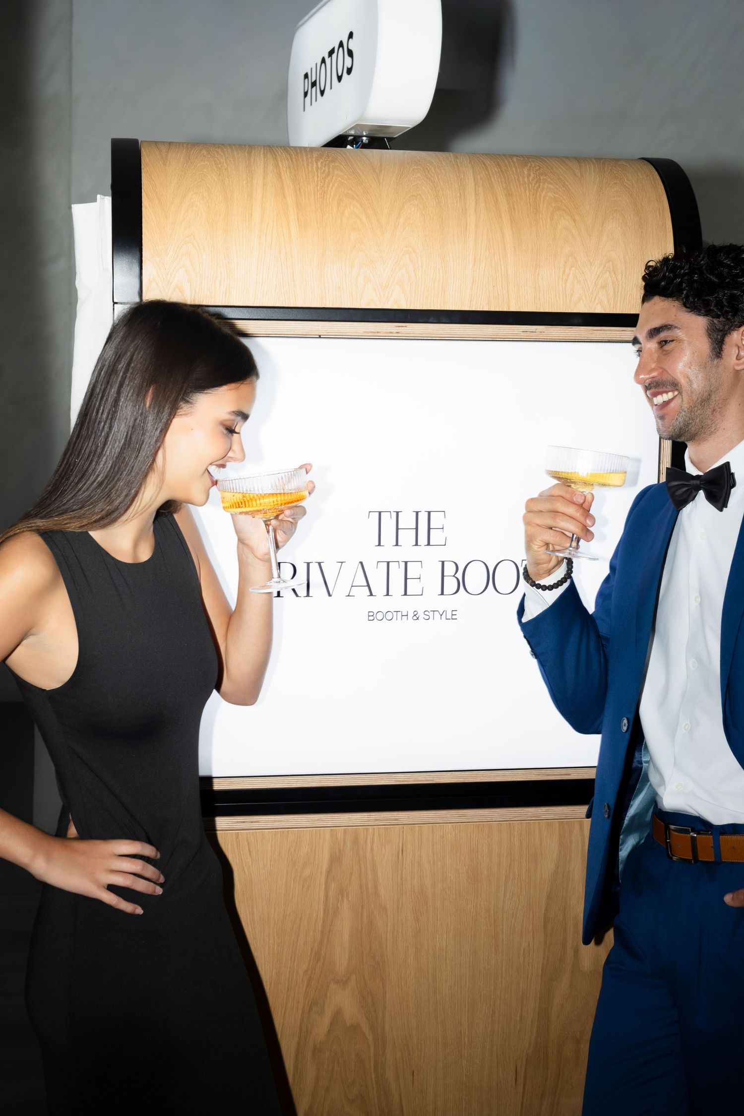 Elegant couple in formal attire toasting cocktails beside The Private Booth photo booth at an upscale event.
