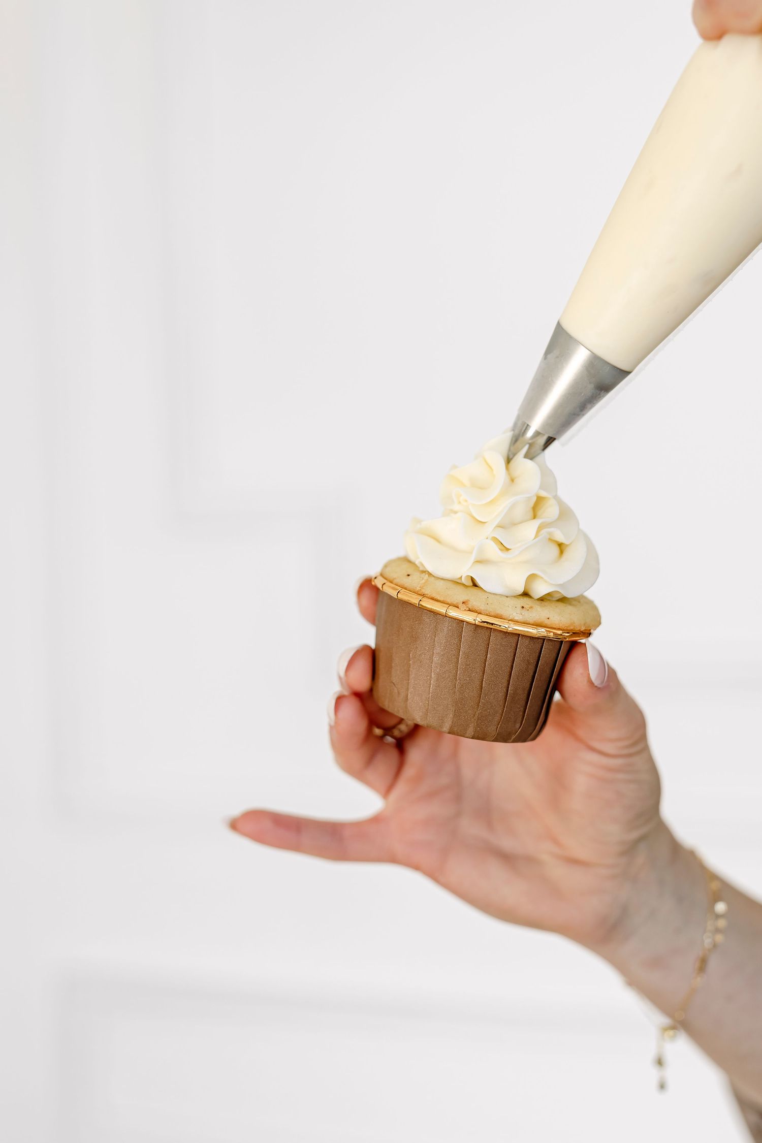 Hand holding a cupcake while piping swirls of white frosting on top with a pastry bag.