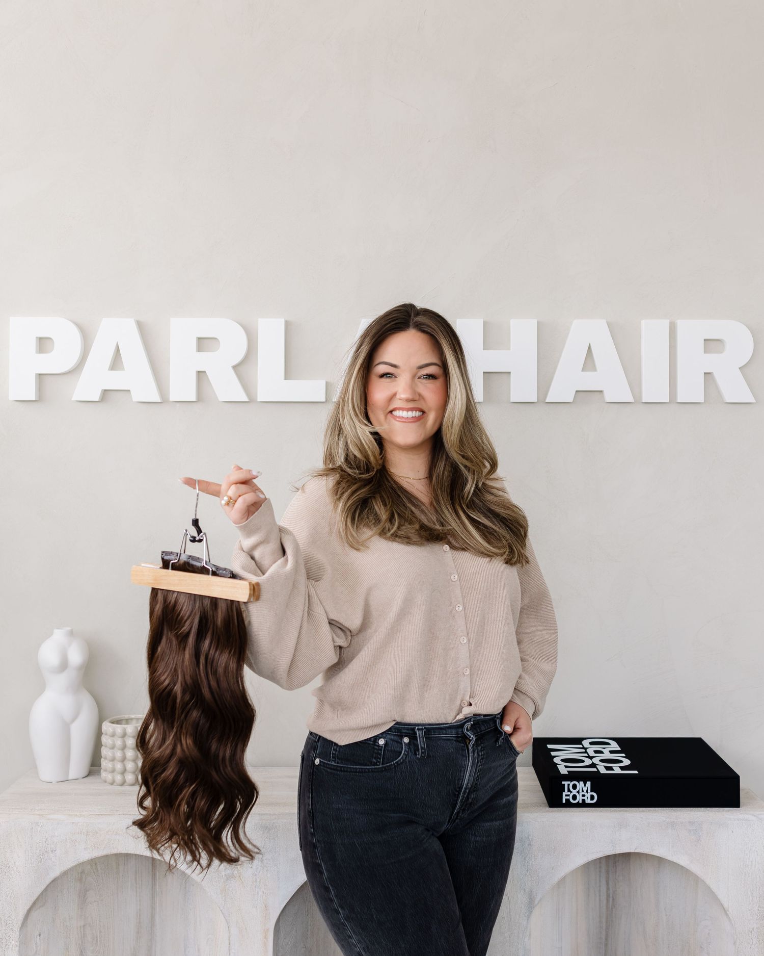 Woman in neutral tones holding hair extensions against white background showing different shades and textures.